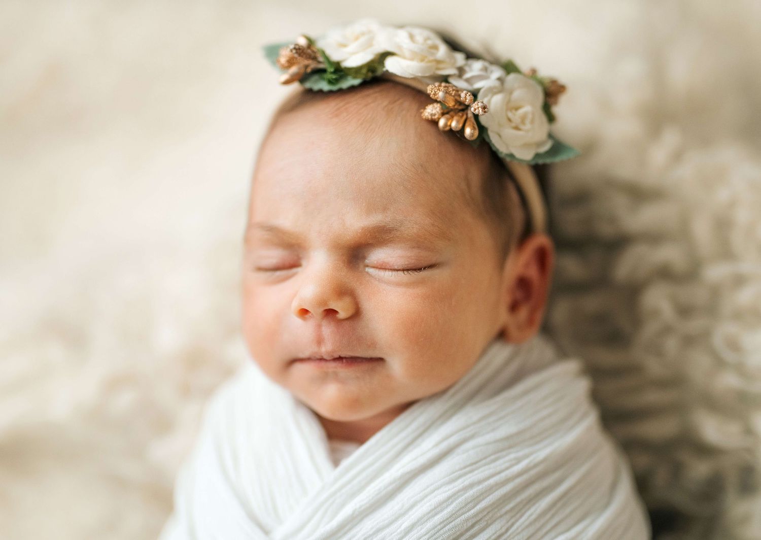 Sleeping newborn baby wrapped in white swaddle wearing a delicate floral headband with white roses.