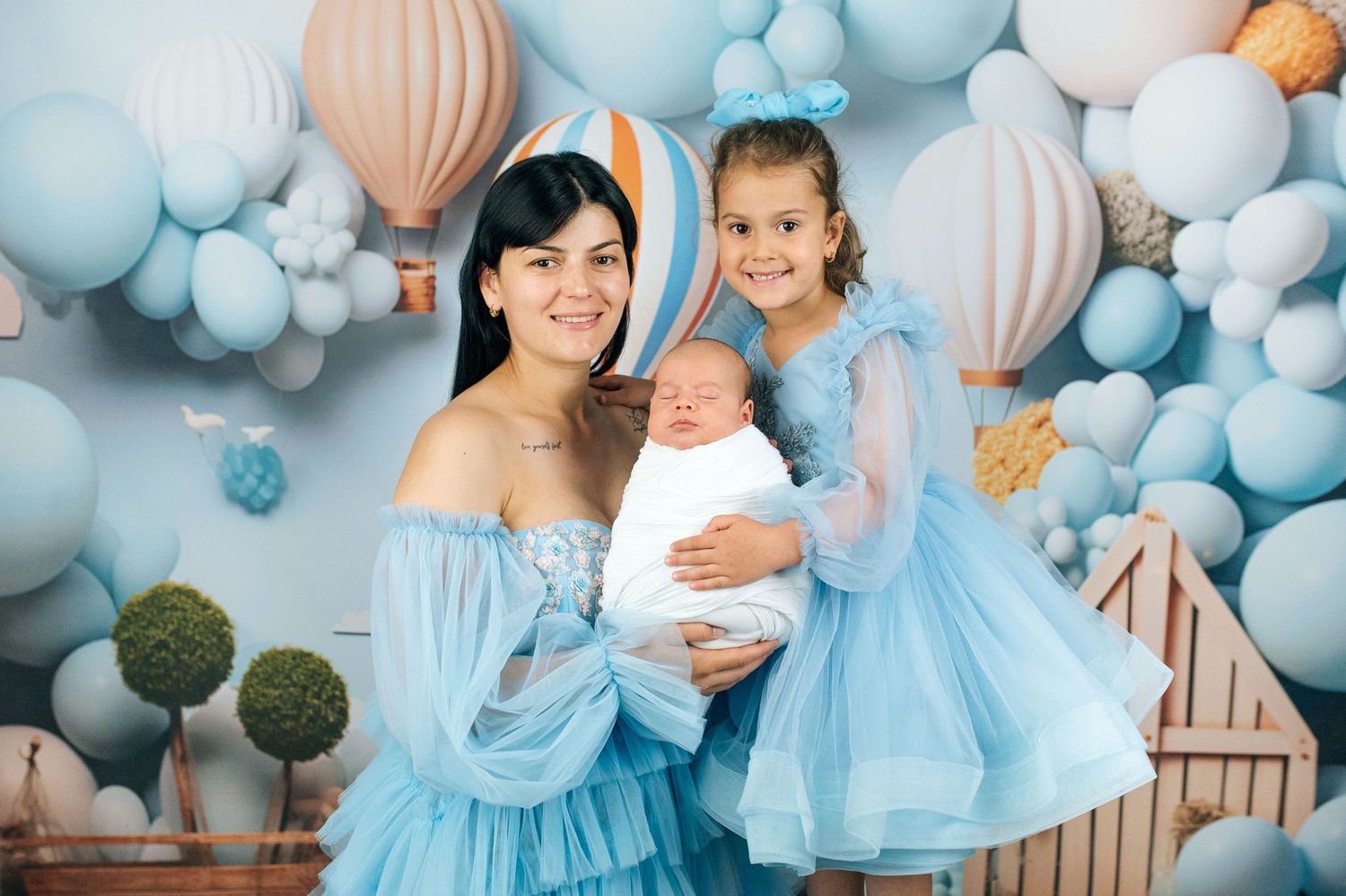 Woman and girl in blue dresses pose with newborn baby against blue balloon backdrop.