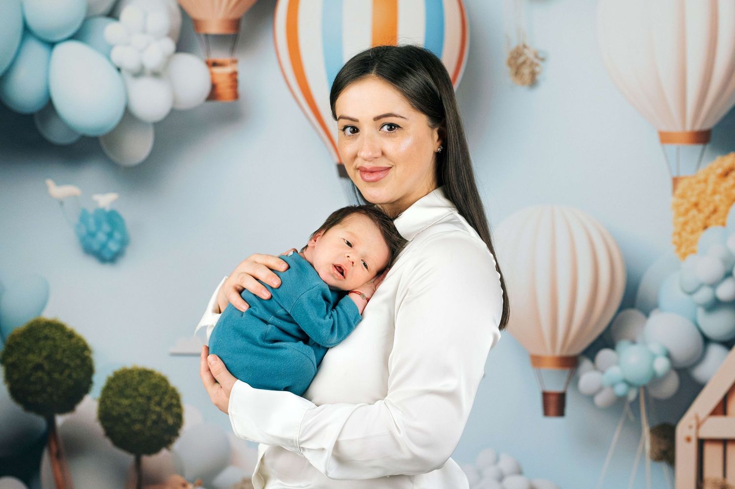 Smiling mother in white outfit holding her newborn baby wrapped in blue blanket with balloon backdrop.