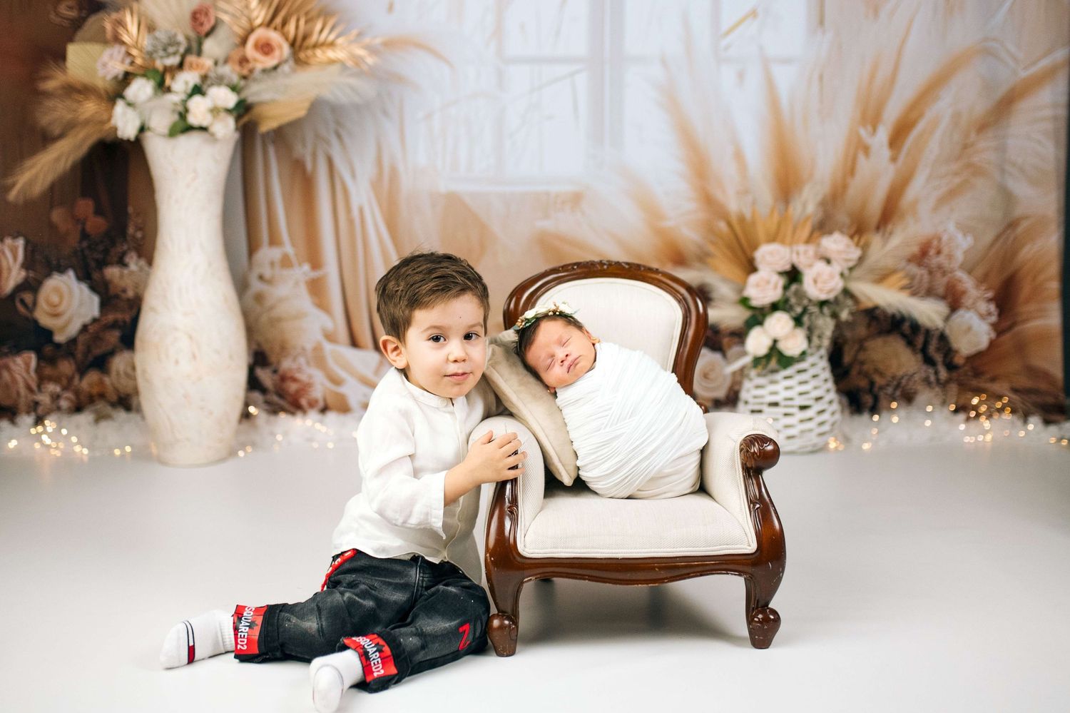A toddler boy sits beside a tiny armchair holding a sleeping newborn baby in a soft floral studio setting.