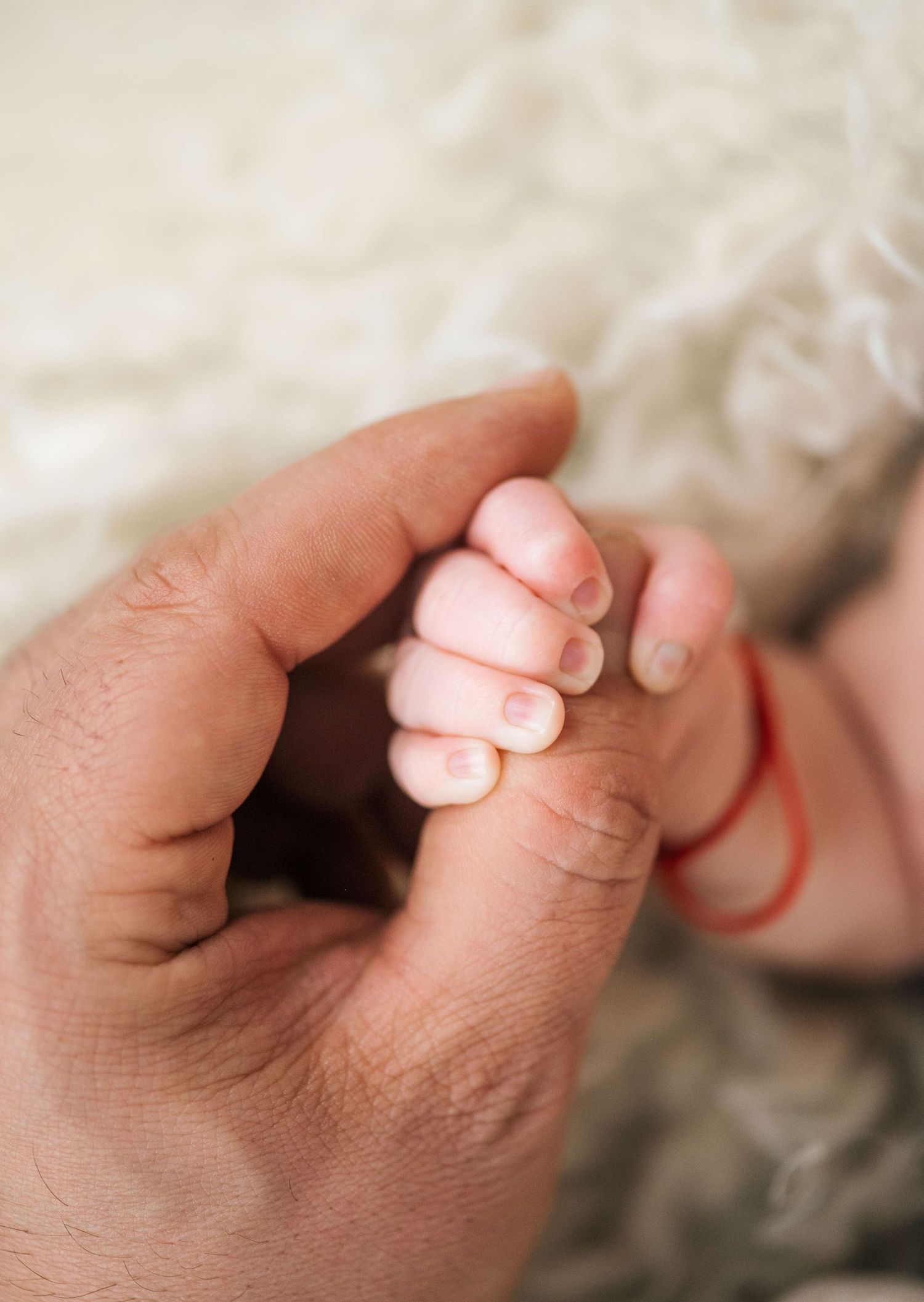 A newborn baby's tiny hand gripping an adult's finger, with a red string bracelet on the wrist.