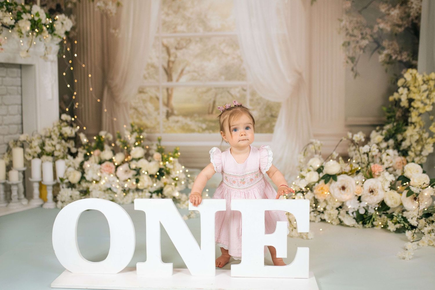 Adorable baby girl in pink dress stands behind large white ONE letters at her first birthday photoshoot.
