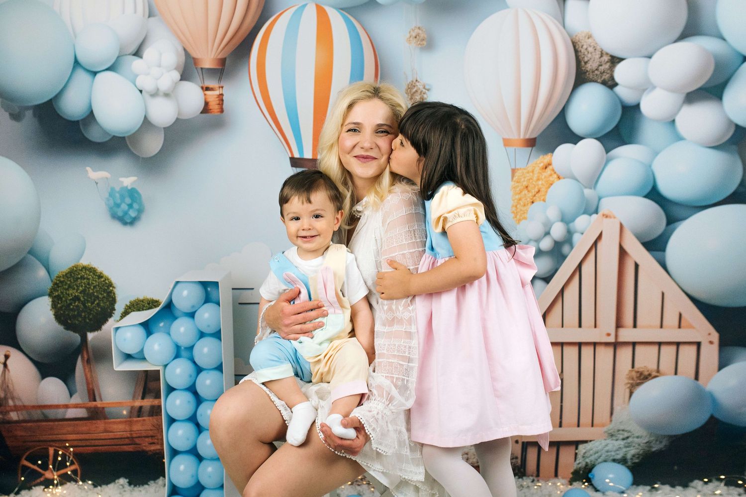 A woman holds a baby while a young girl kisses her cheek, surrounded by blue balloons and hot air balloon decorations.
