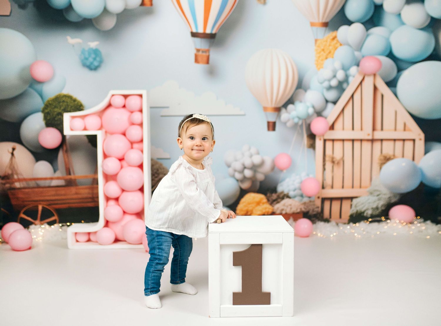 Toddler smiling at first birthday photo shoot with balloons, hot air balloons backdrop, and number 1 blocks.