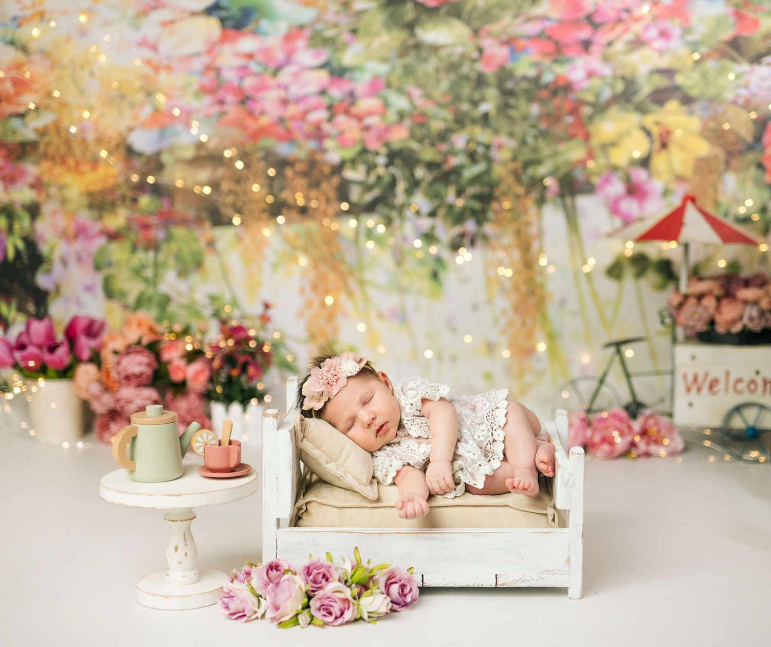 Newborn baby sleeping peacefully on a white miniature bed surrounded by flowers and fairy lights in a garden-themed photo setup.