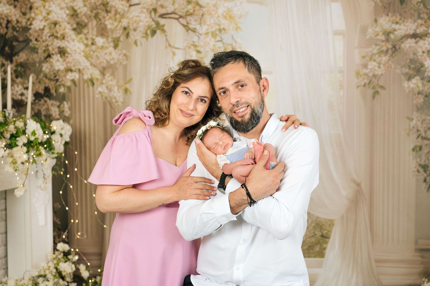 Happy couple in elegant attire holding newborn baby in floral decorated studio setting.