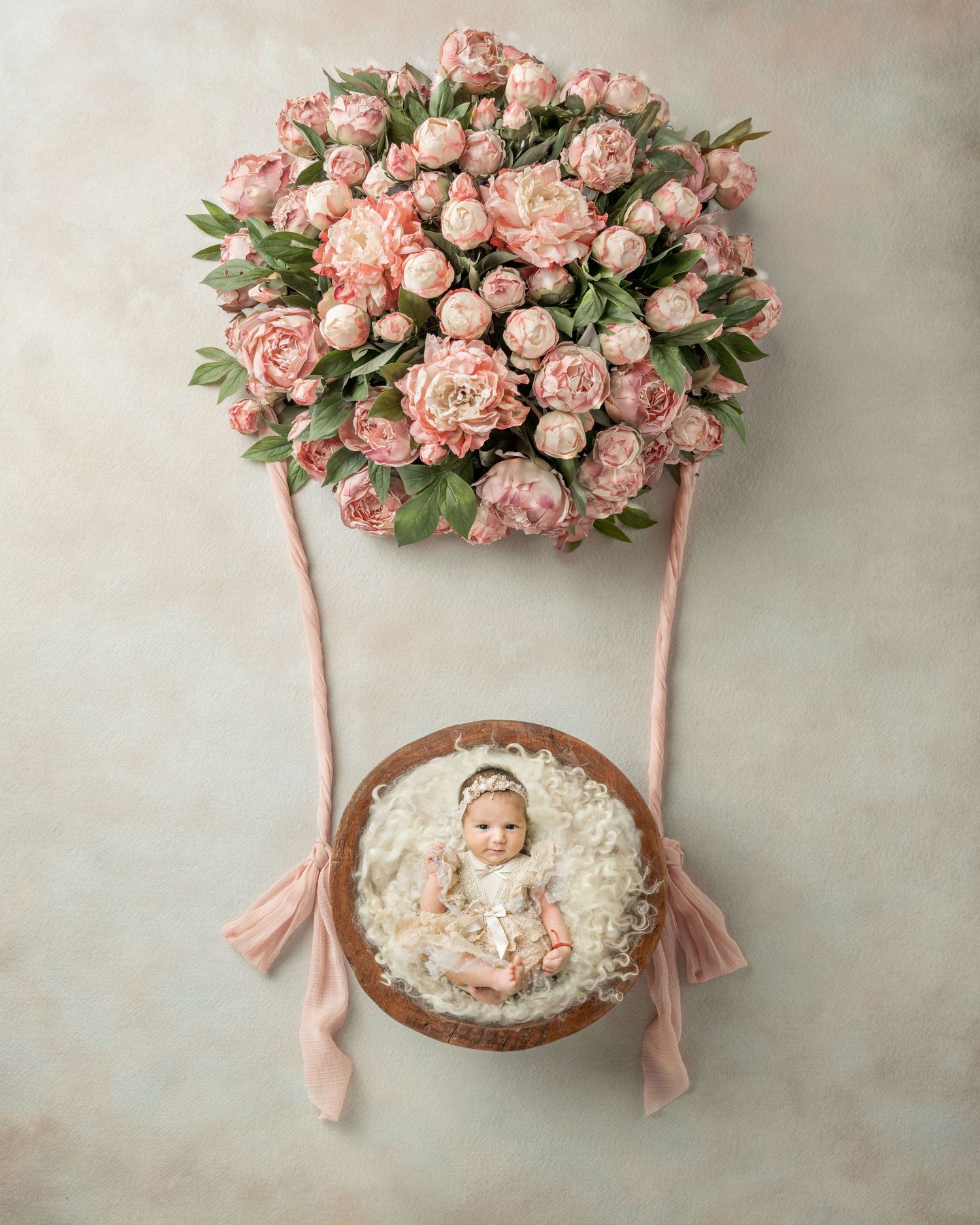 Newborn baby in a wooden bowl beneath a pink peony hot air balloon arrangement on a soft pastel background.