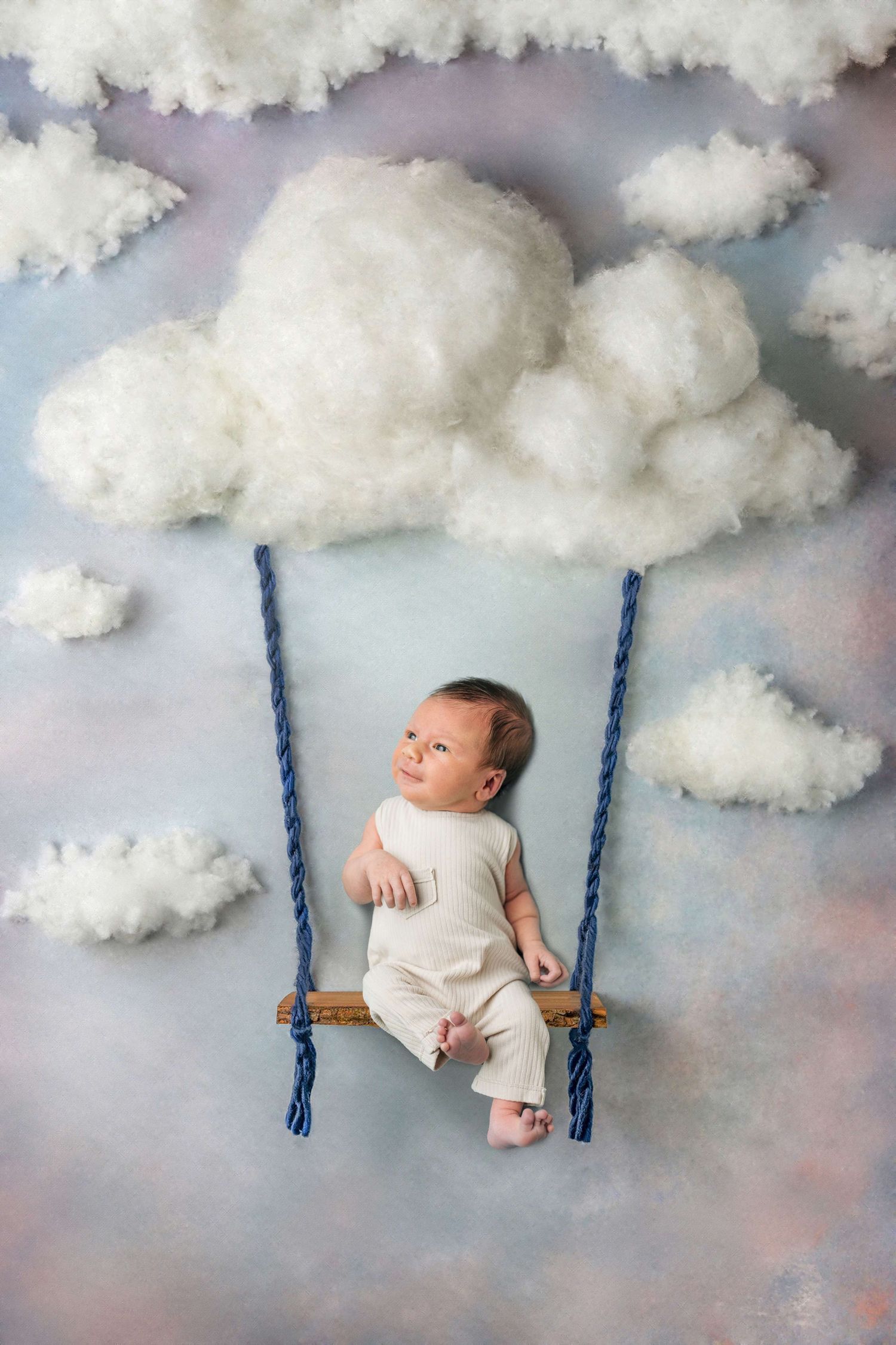 Baby in white outfit lying on a wooden swing surrounded by fluffy white clouds on a dreamy blue sky backdrop.