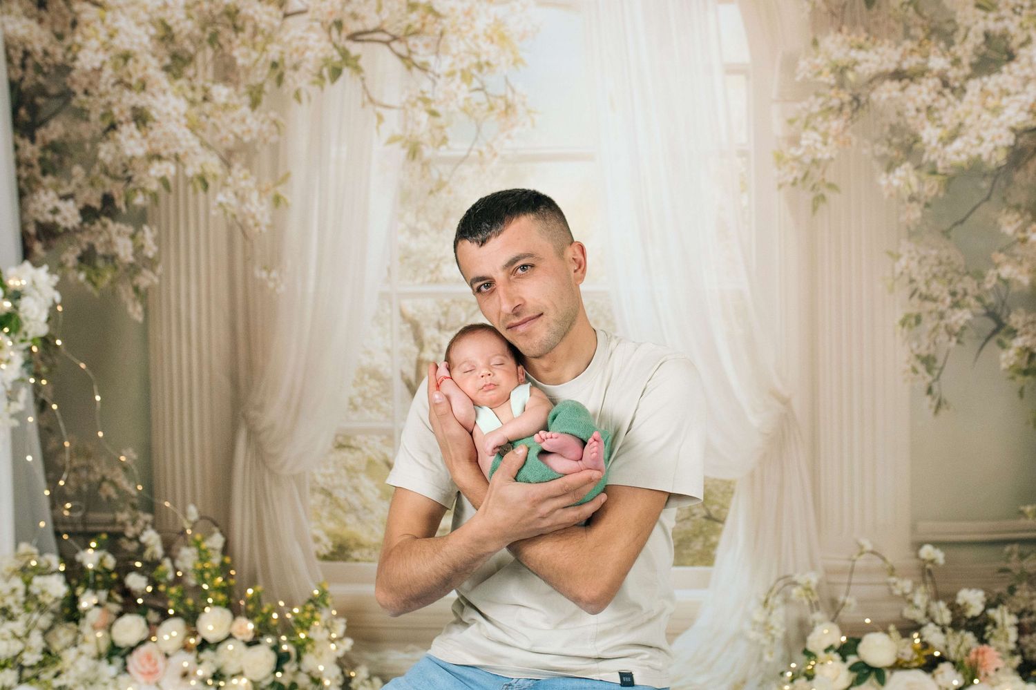 Father holding sleeping newborn baby surrounded by white floral decorations in a professional photo studio setting.