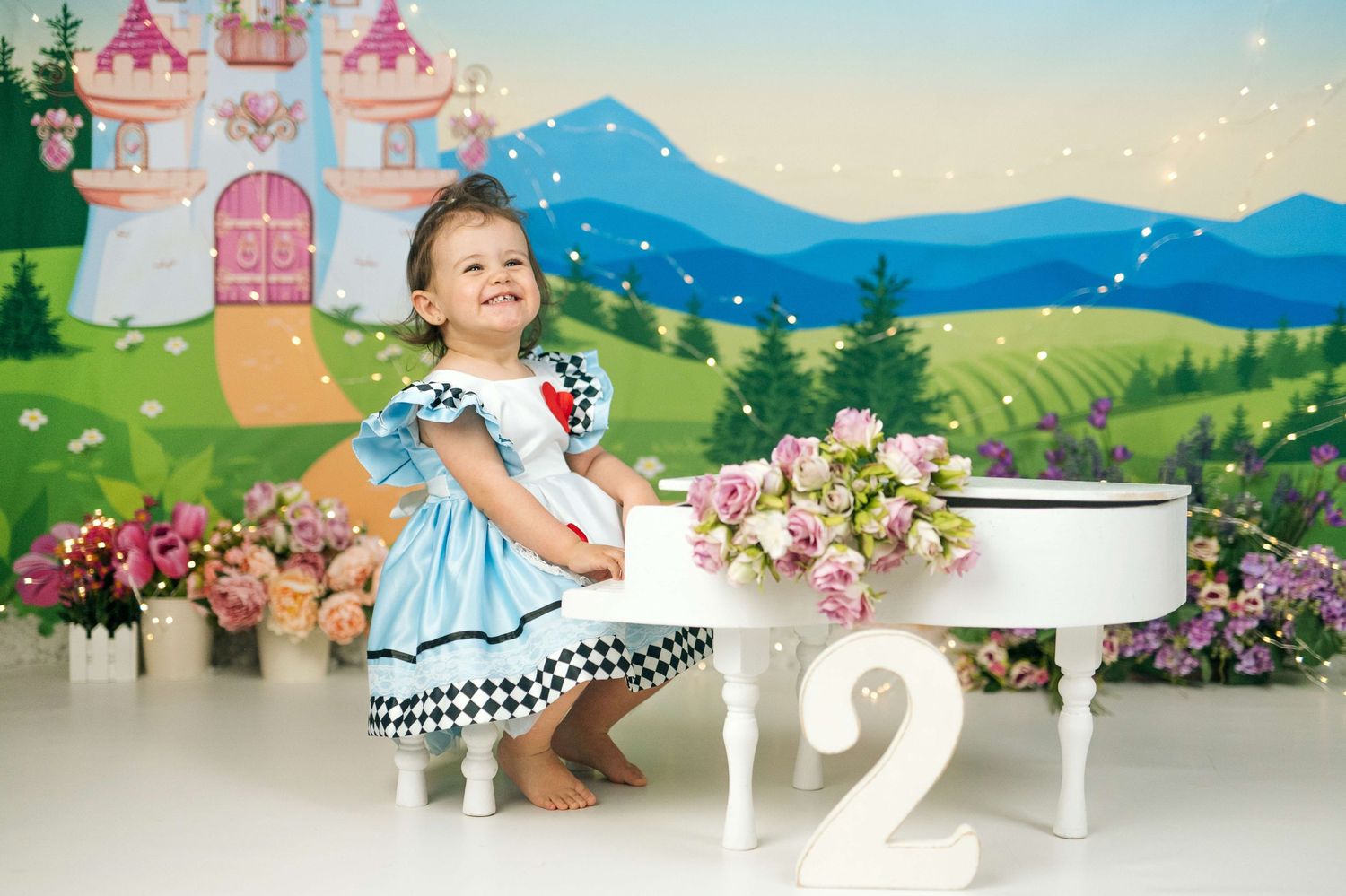 Toddler girl in Alice in Wonderland dress poses near white piano and number 2 with floral fairy tale castle backdrop.