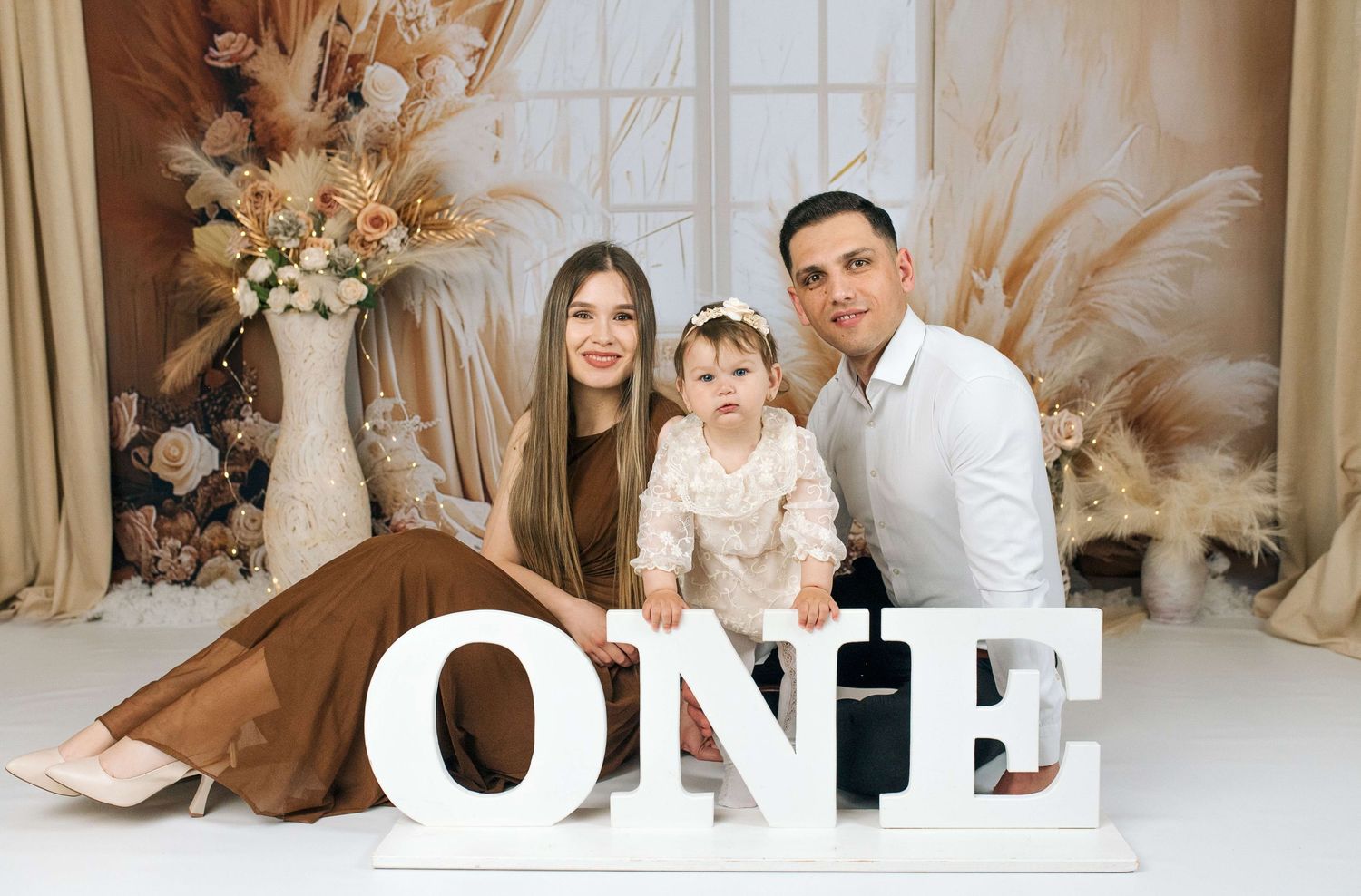 Family of three posing with large ONE sign for baby first birthday photoshoot with boho floral backdrop.