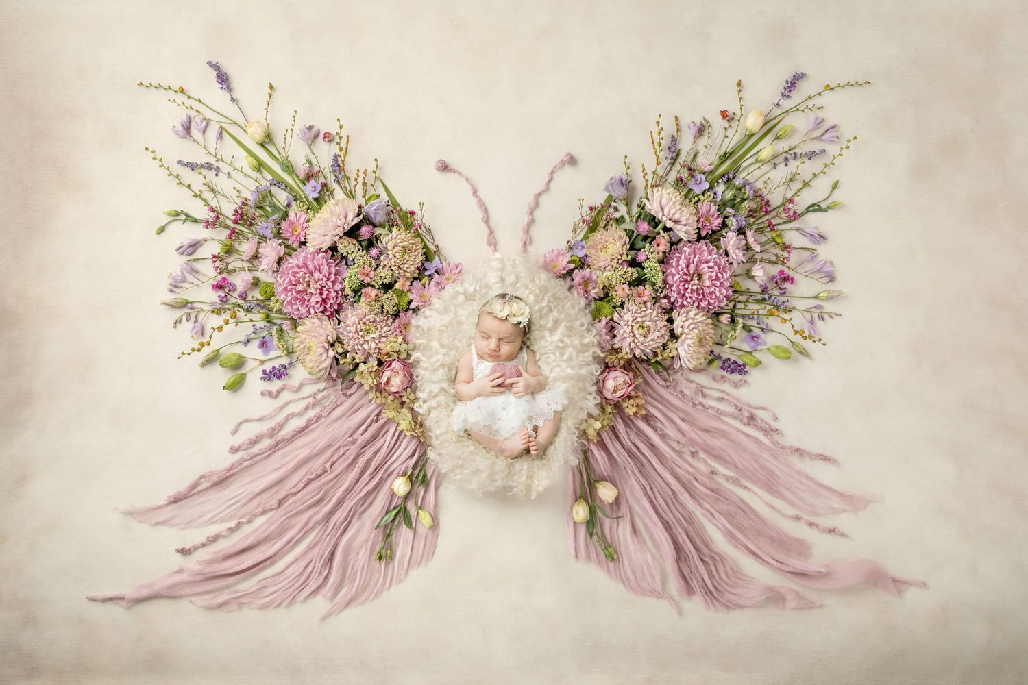 Newborn baby sleeping in floral butterfly wings arrangement with pink fabric and colorful spring flowers.