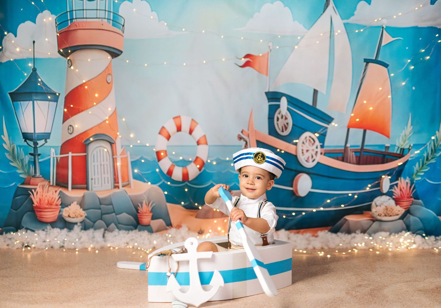 Baby in sailor outfit sits in anchor box with nautical backdrop featuring lighthouse, sailboat, and ocean decorations.