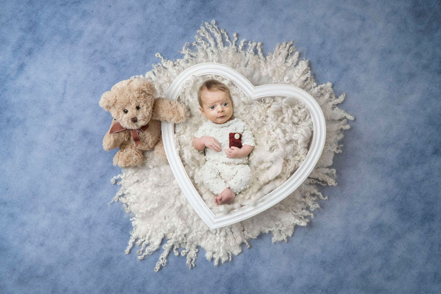 Newborn baby sleeping in white heart-shaped prop with teddy bear on blue background.