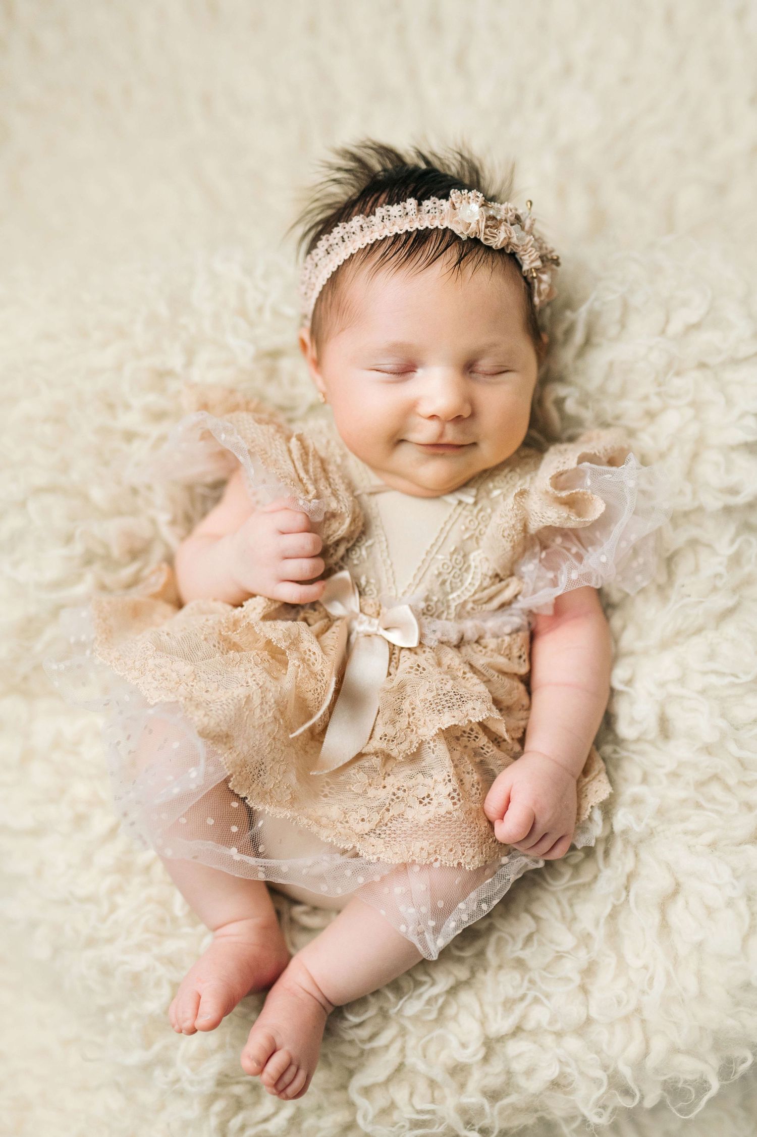Smiling newborn baby girl in beige lace dress and floral headband lying on fluffy cream blanket.
