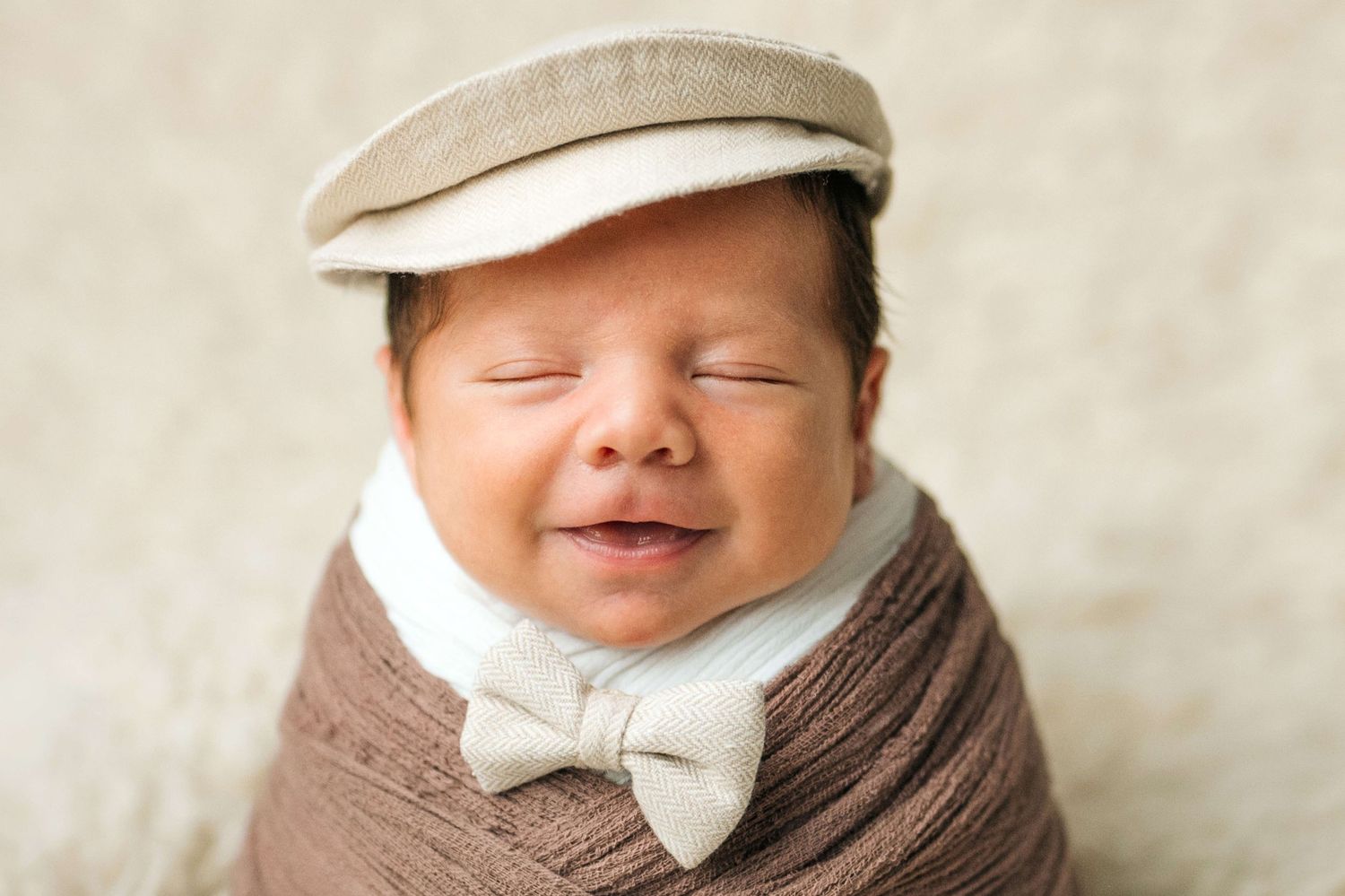 Smiling newborn baby wrapped in brown swaddle wearing a beige flat cap and bow tie for a professional photo shoot.