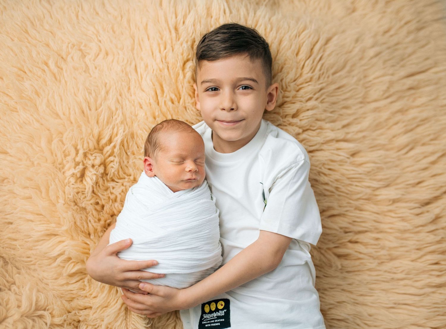 Older boy in white shirt gently holds swaddled newborn baby sibling on fluffy beige fur backdrop.
