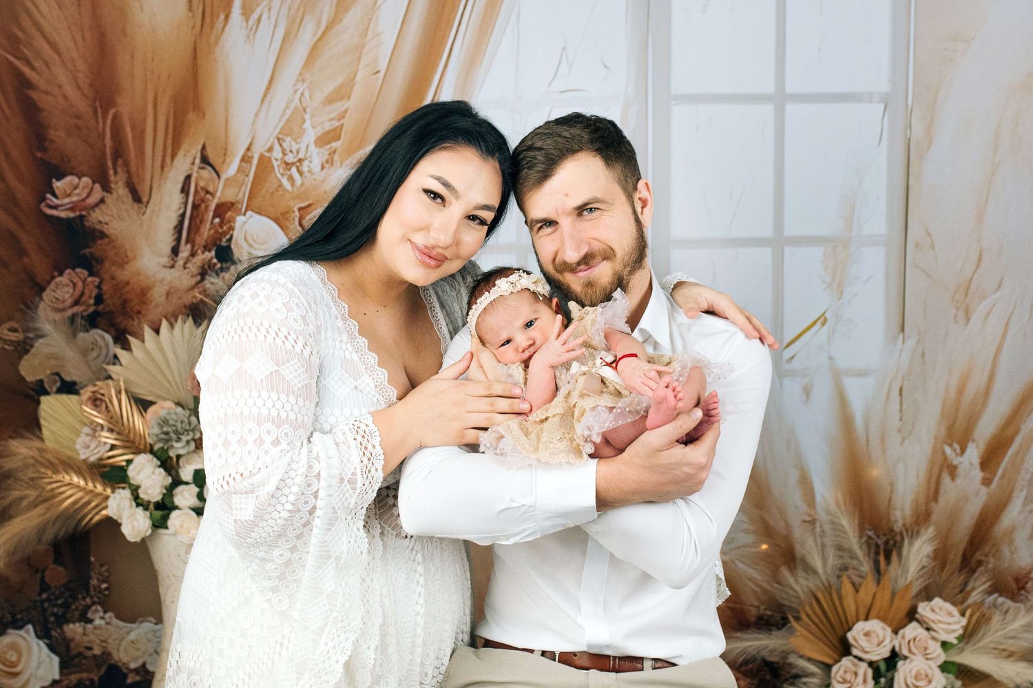 A happy couple in elegant white attire holds their newborn baby surrounded by boho floral decor.