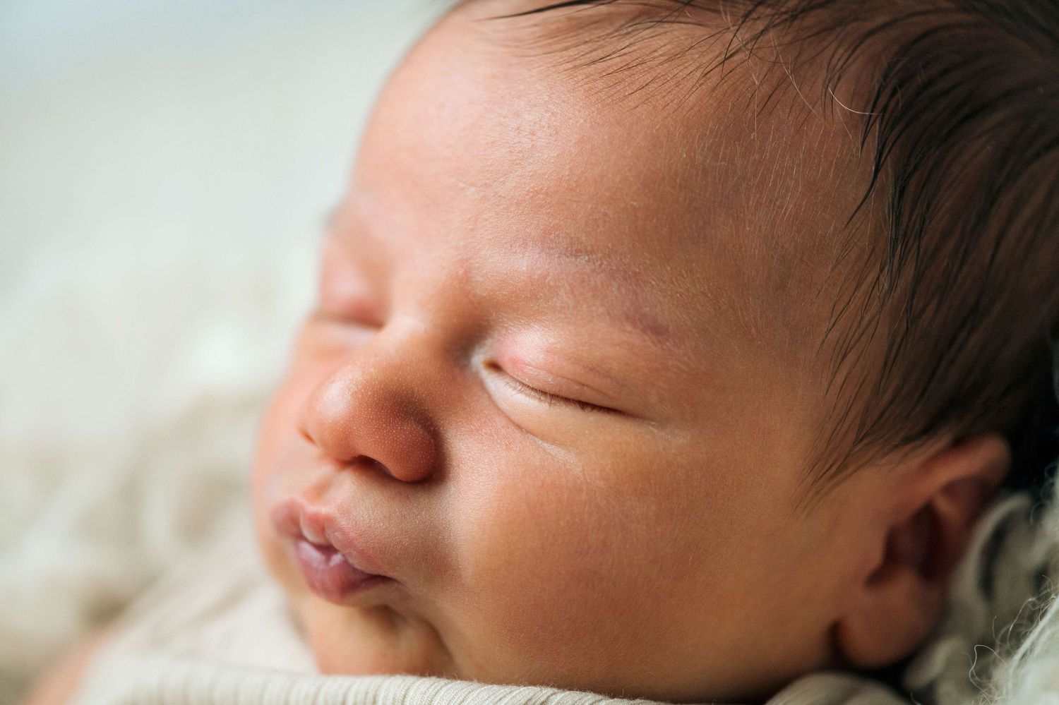 Close-up portrait of a sleeping newborn baby with dark hair, peaceful expression on a soft cream background.
