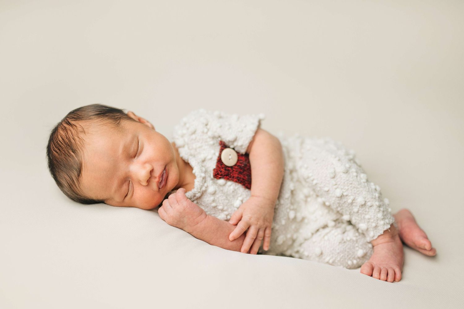 Sleeping newborn baby in white textured romper with red bow tie on cream background.