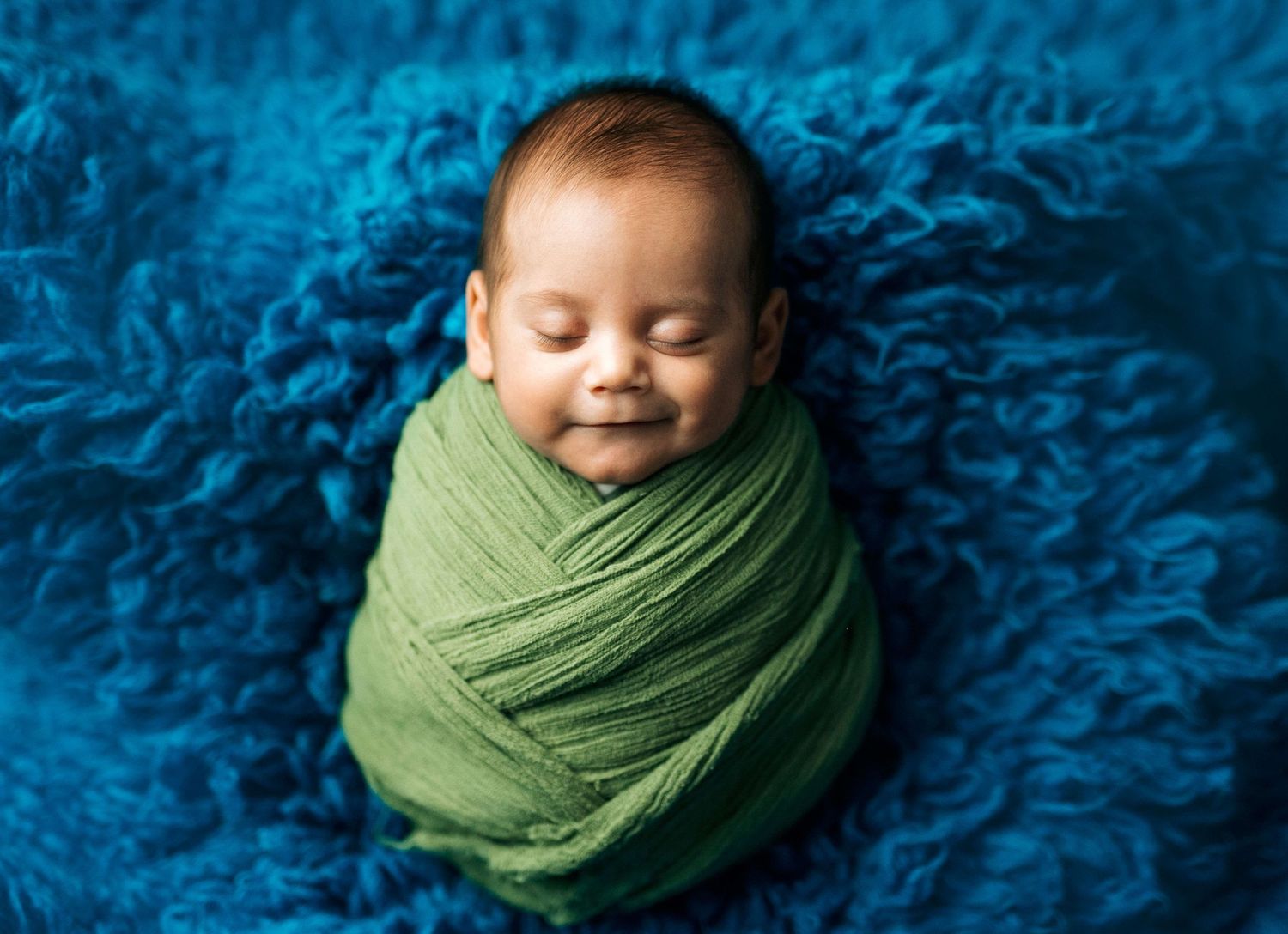 Smiling newborn baby swaddled in green wrap sleeping on blue fluffy blanket in professional portrait photo.