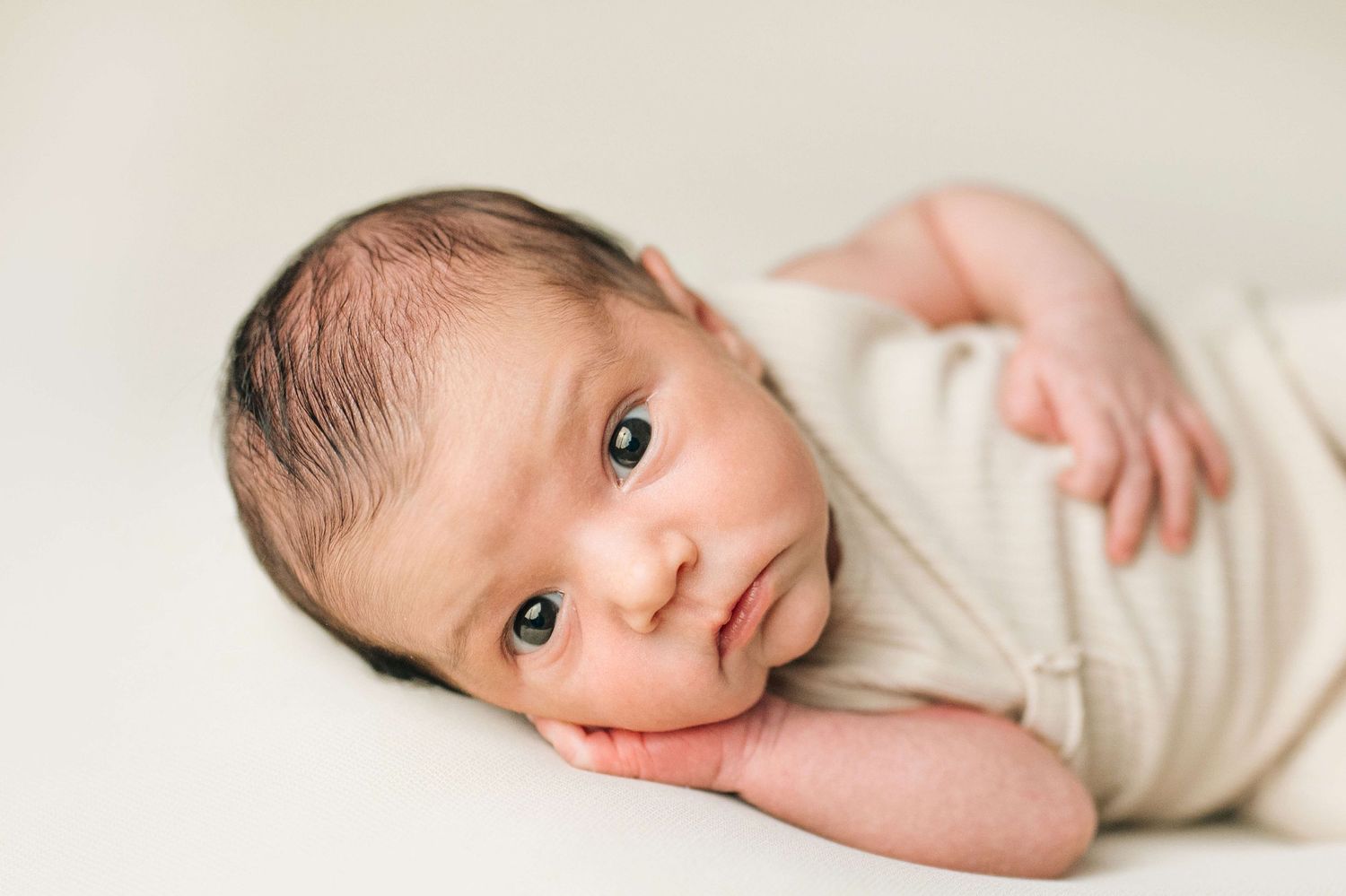 Newborn baby wrapped in cream swaddle lying on white background, looking at camera with dark eyes.