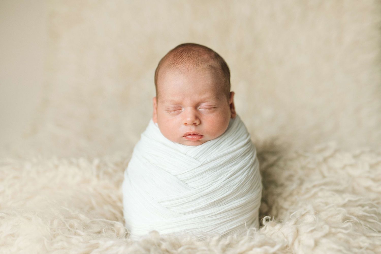 Peacefully sleeping newborn baby swaddled in a soft white wrap, posed on a fluffy cream fur blanket.