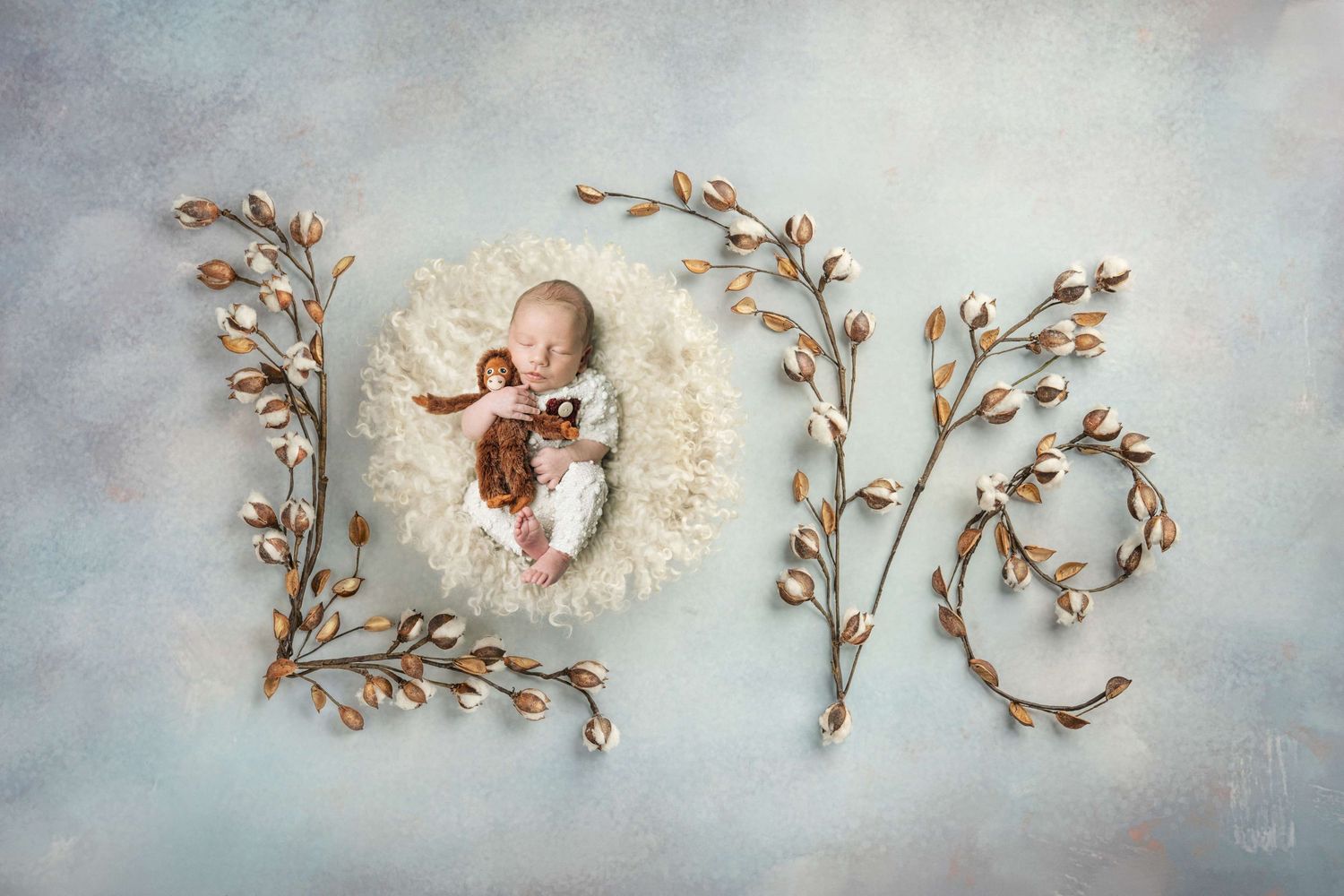 Newborn baby sleeping in fluffy white bowl surrounded by golden floral branches spelling LOVE on soft blue background.