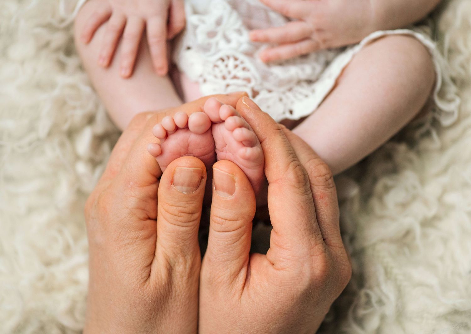 Adult hands gently cradling tiny newborn baby feet on a soft white fluffy blanket.