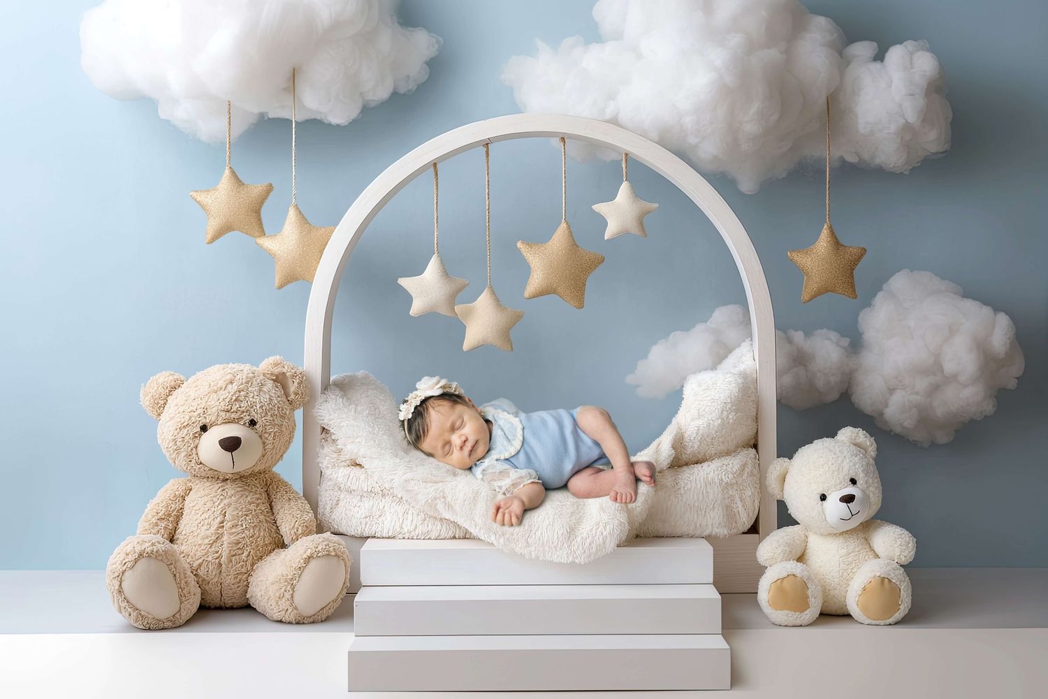 Newborn baby sleeping in a dreamy white arch bed surrounded by teddy bears, fluffy clouds, and gold stars on blue backdrop.