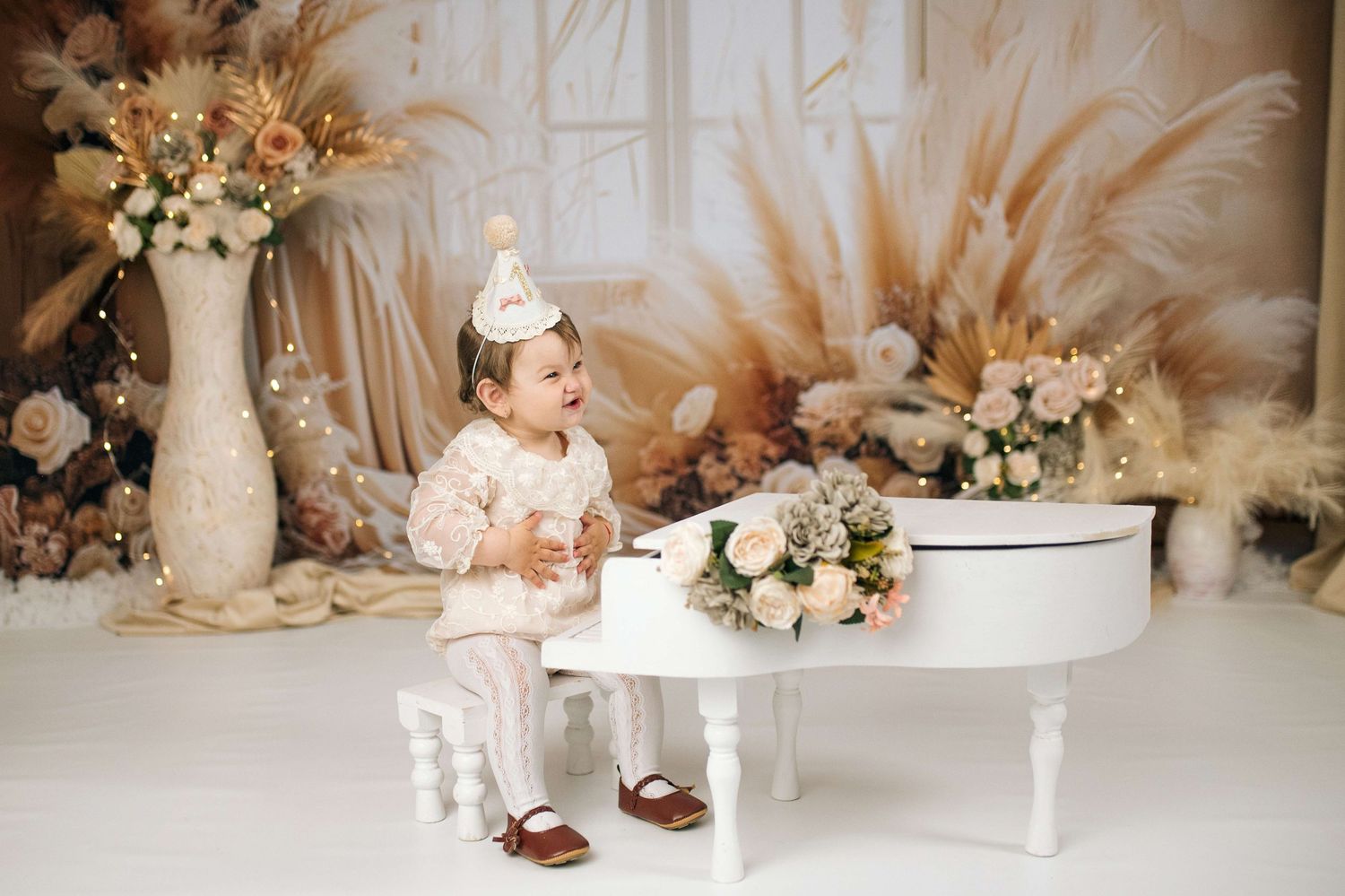 A joyful toddler in a party hat sits beside a white piano surrounded by elegant floral decor in a warm, dreamy setting.