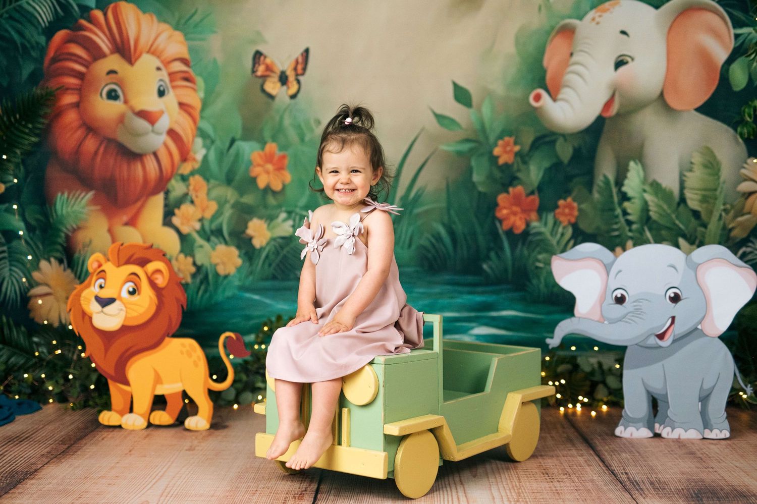 A smiling girl in a pink dress sits on a toy jeep in front of a colorful jungle backdrop with cartoon lions and elephants.