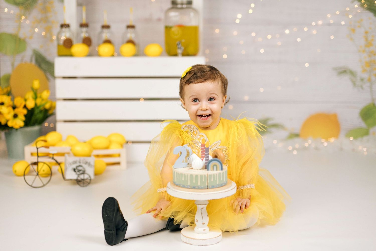 Smiling toddler in yellow tutu dress sits beside a mint birthday cake in a lemon-themed photo studio setting.