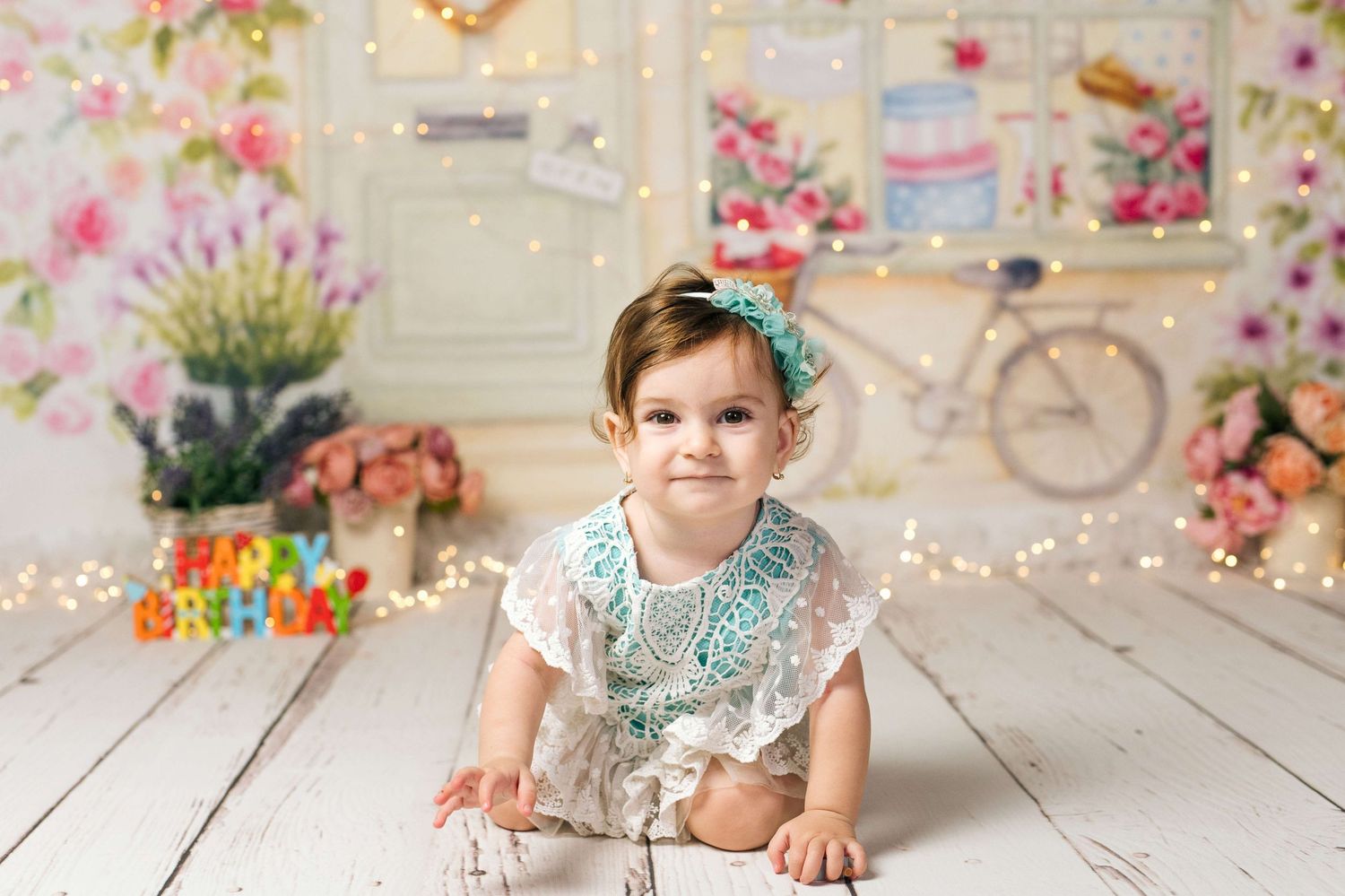 Adorable baby girl in lace outfit crawling on white wood floor with floral birthday backdrop and fairy lights.
