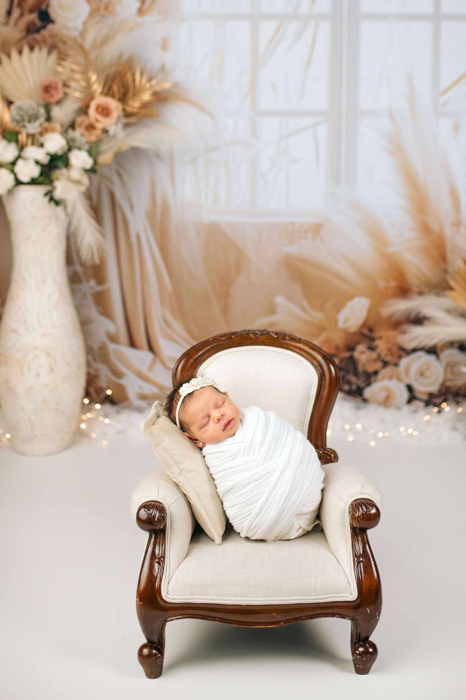 Newborn baby swaddled in white wrap sleeping peacefully on a miniature cream armchair with floral decor backdrop.
