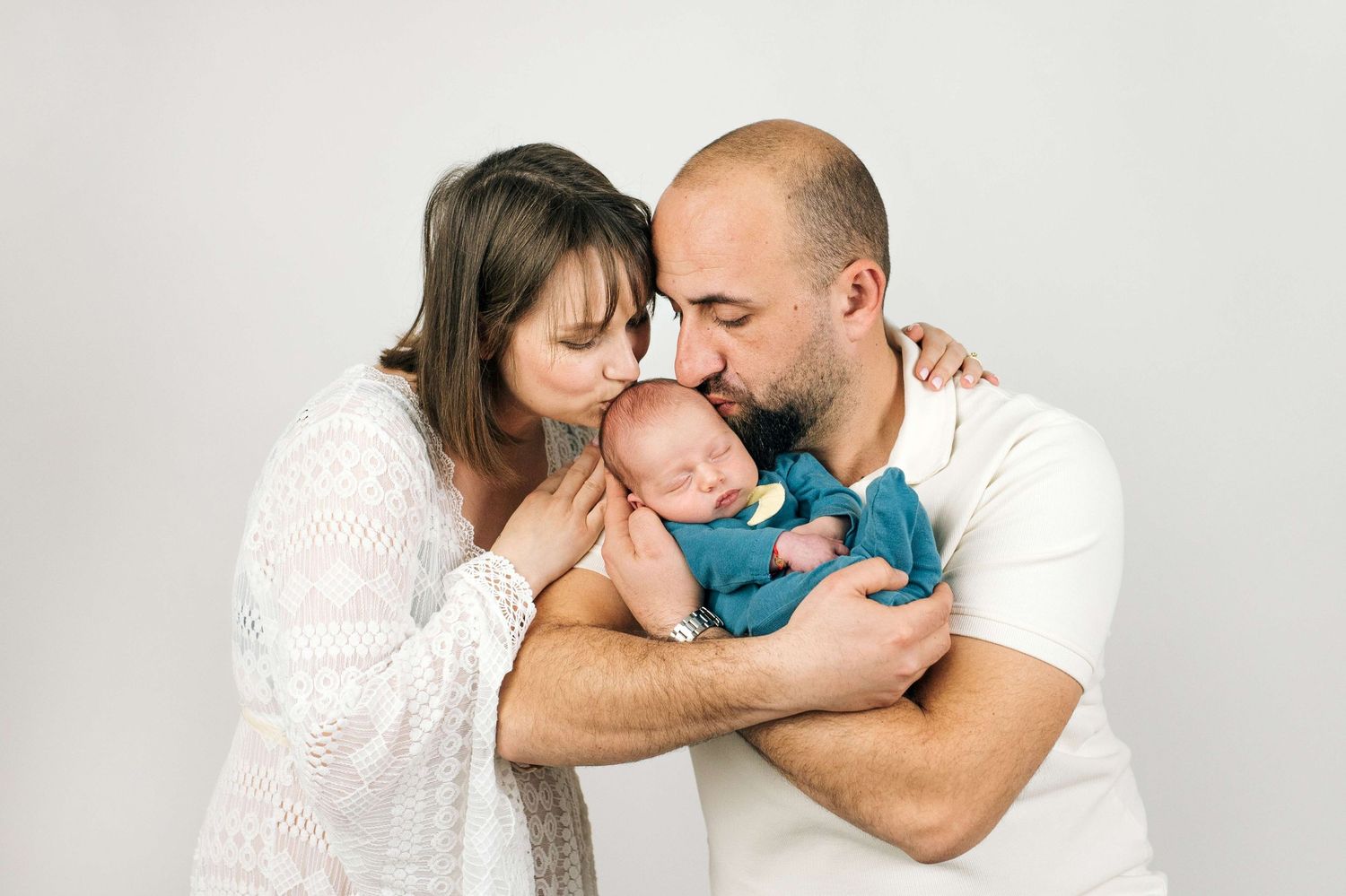 Parents kissing their sleeping newborn baby wrapped in a teal blanket against a white studio background.