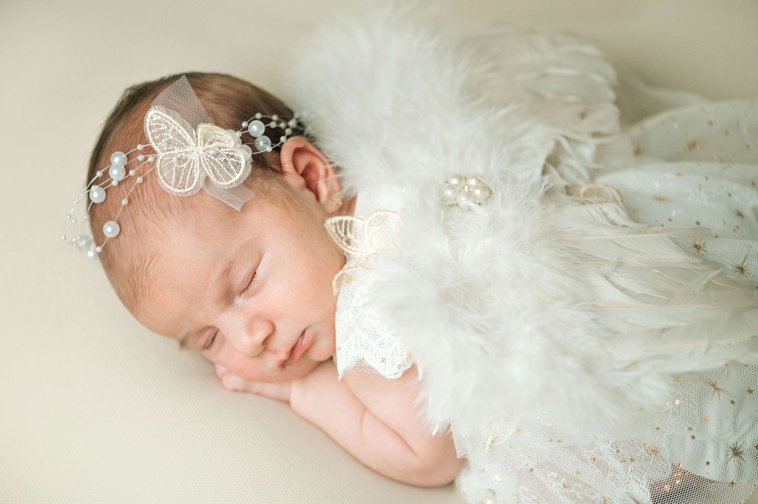 Newborn baby sleeping peacefully wearing white feather wings and a delicate butterfly headband with pearls.