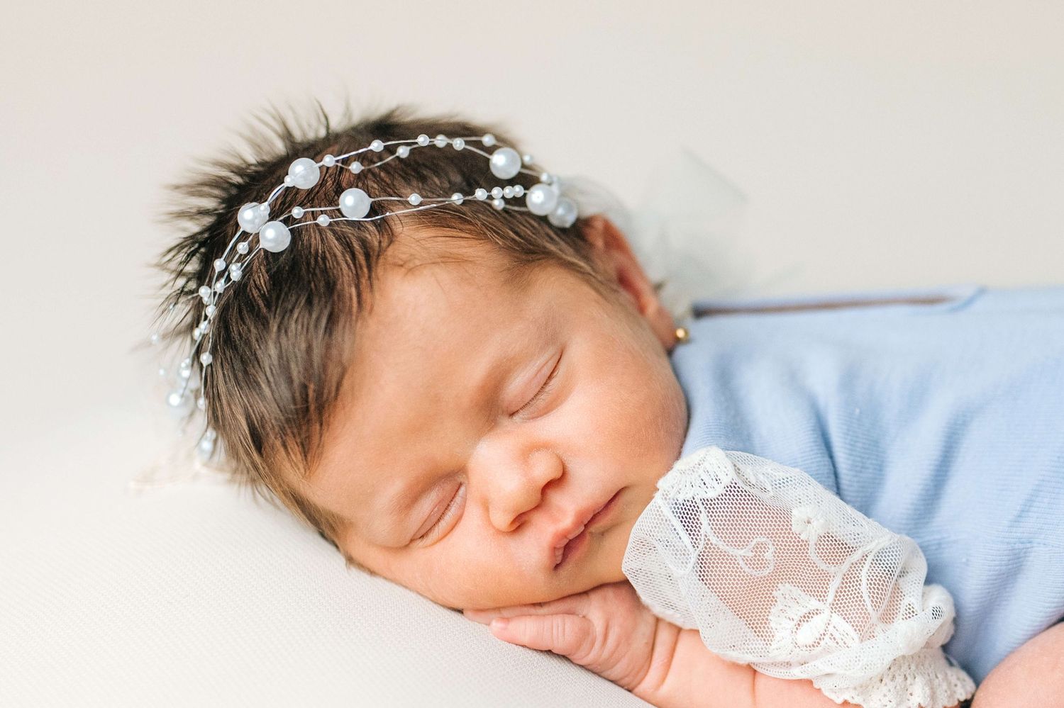 Sleeping newborn baby wearing a floral crown headband and blue wrap, peacefully resting on a white background.