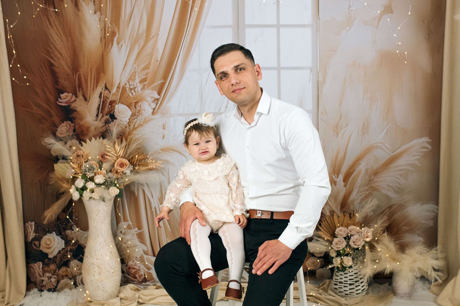 Man in white shirt sits with baby girl in cream dress amid elegant floral backdrop with pampas grass decor.