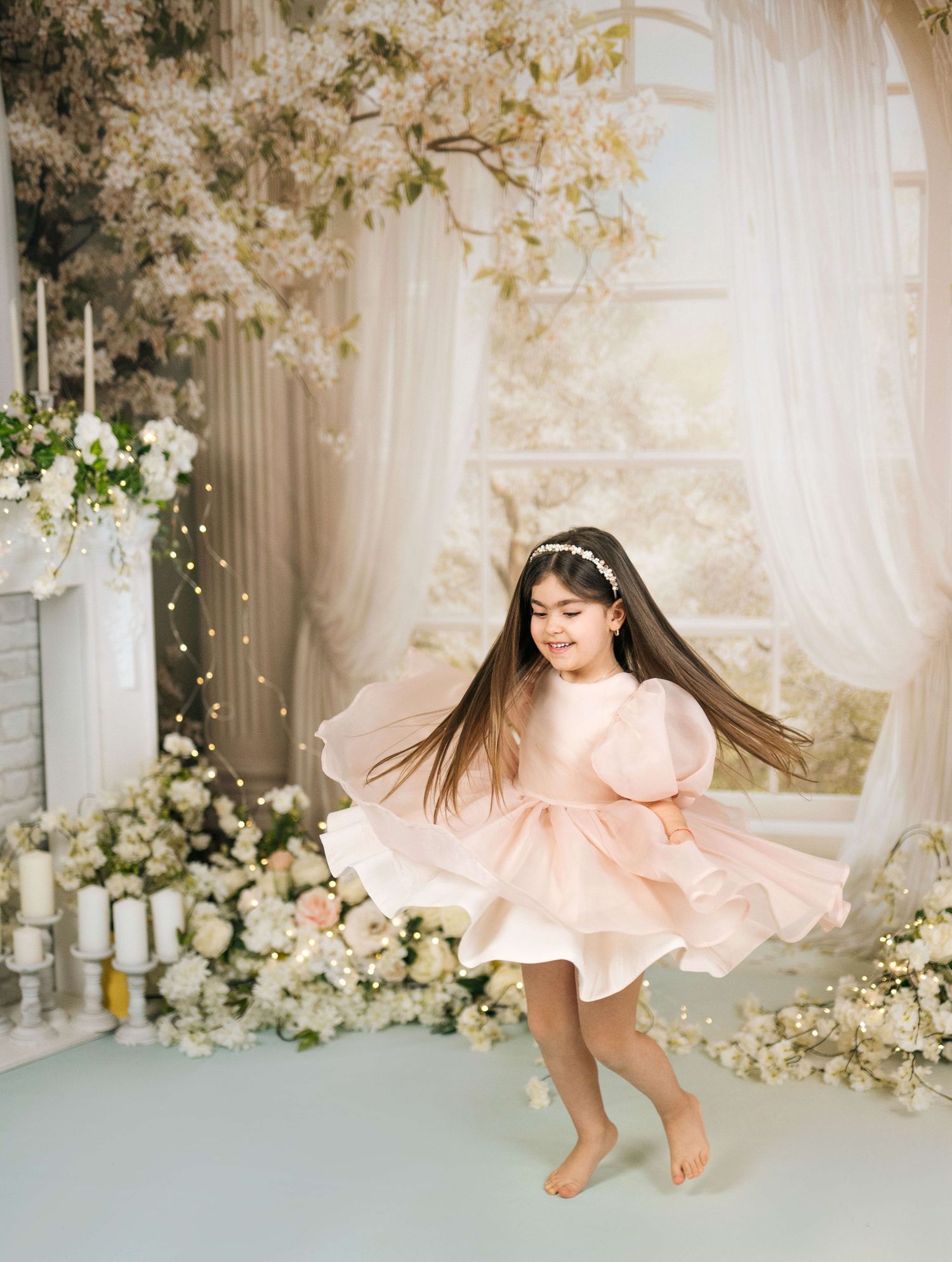 Young girl in pink dress twirling among white floral decorations and sheer curtains in elegant studio setting.