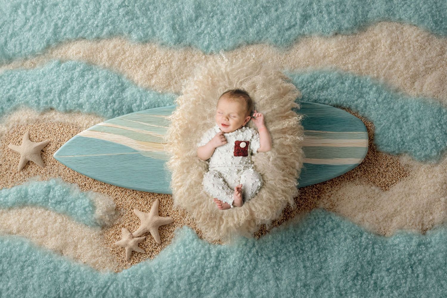 Newborn baby sleeping on a miniature surfboard with starfish and sand on a teal wave backdrop.