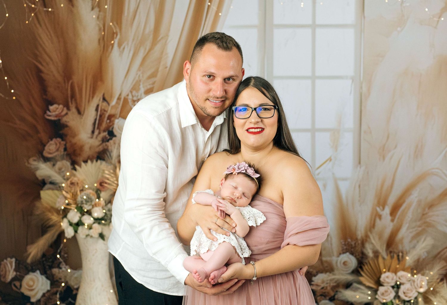 A couple poses with their newborn baby in a boho-styled photo shoot with floral and pampas grass decor.