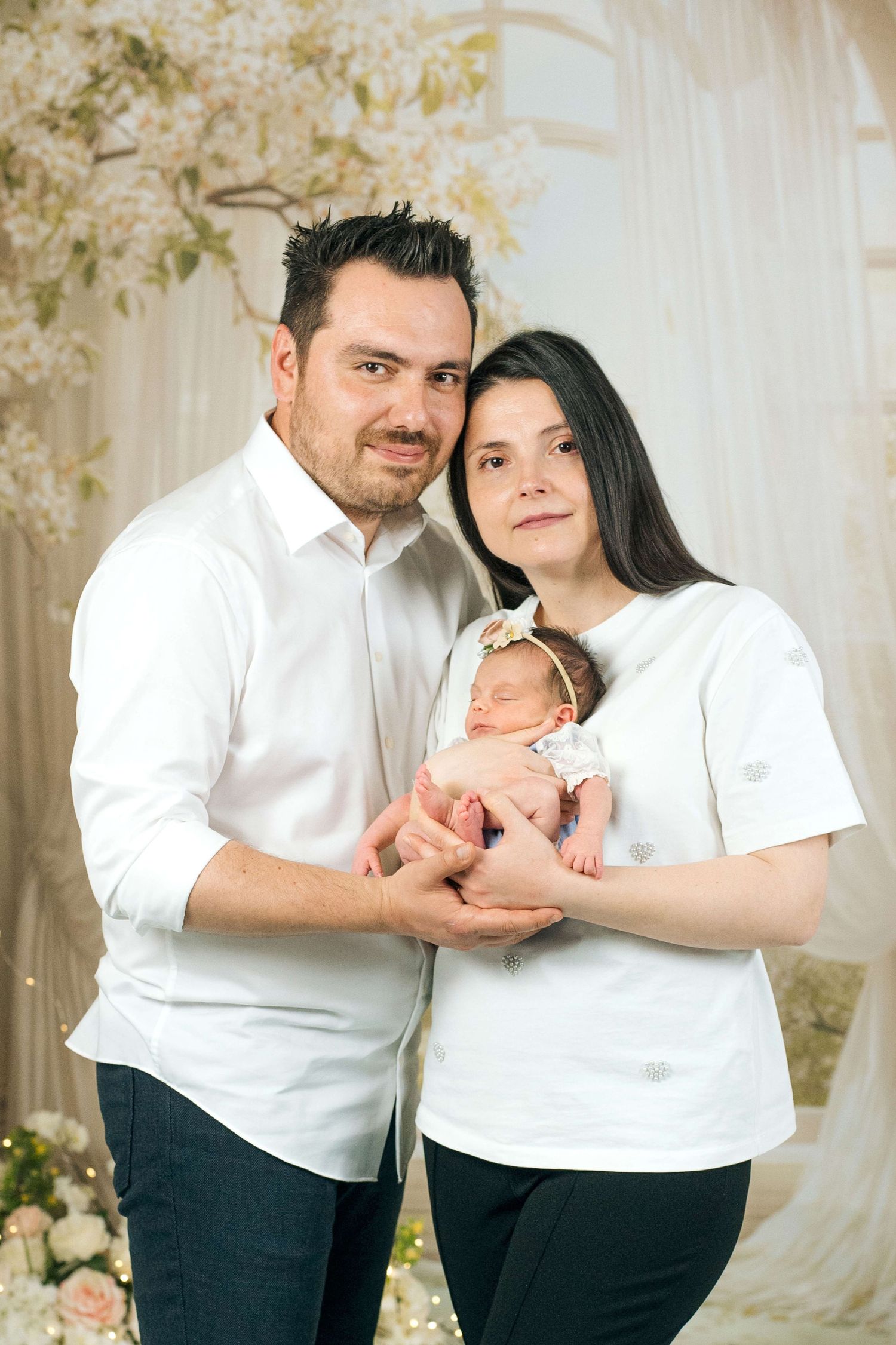 Happy parents in white shirts holding their newborn baby girl with a floral headband in a studio setting.