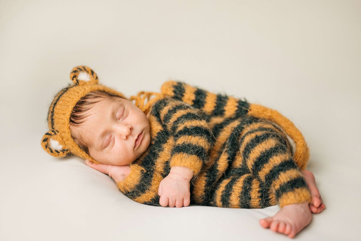 Sleeping newborn baby wearing a knitted tiger striped onesie and matching bear ear hat on white background.