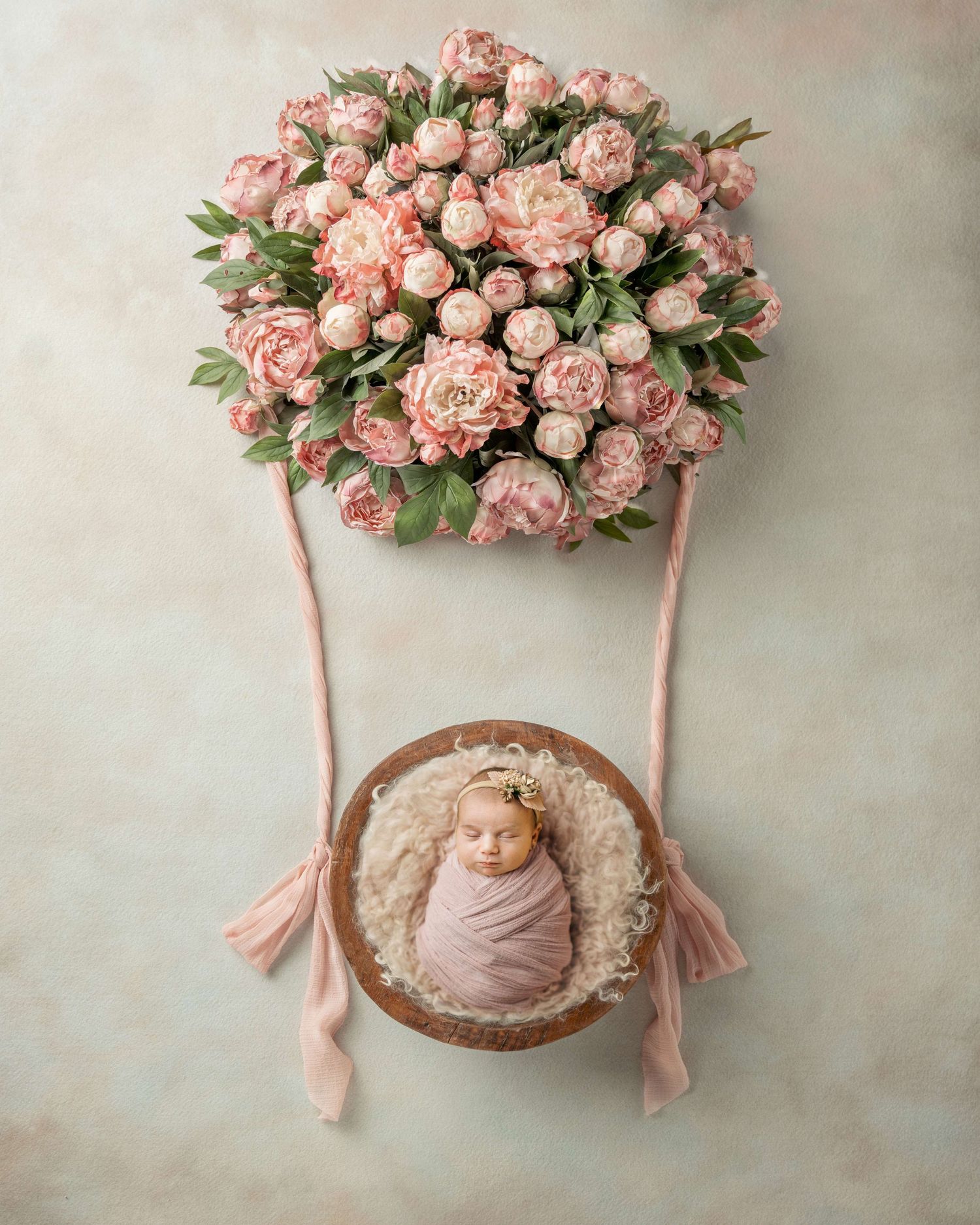 Newborn baby wrapped in pink swaddled in a wooden bowl beneath a hot air balloon of pink peonies.