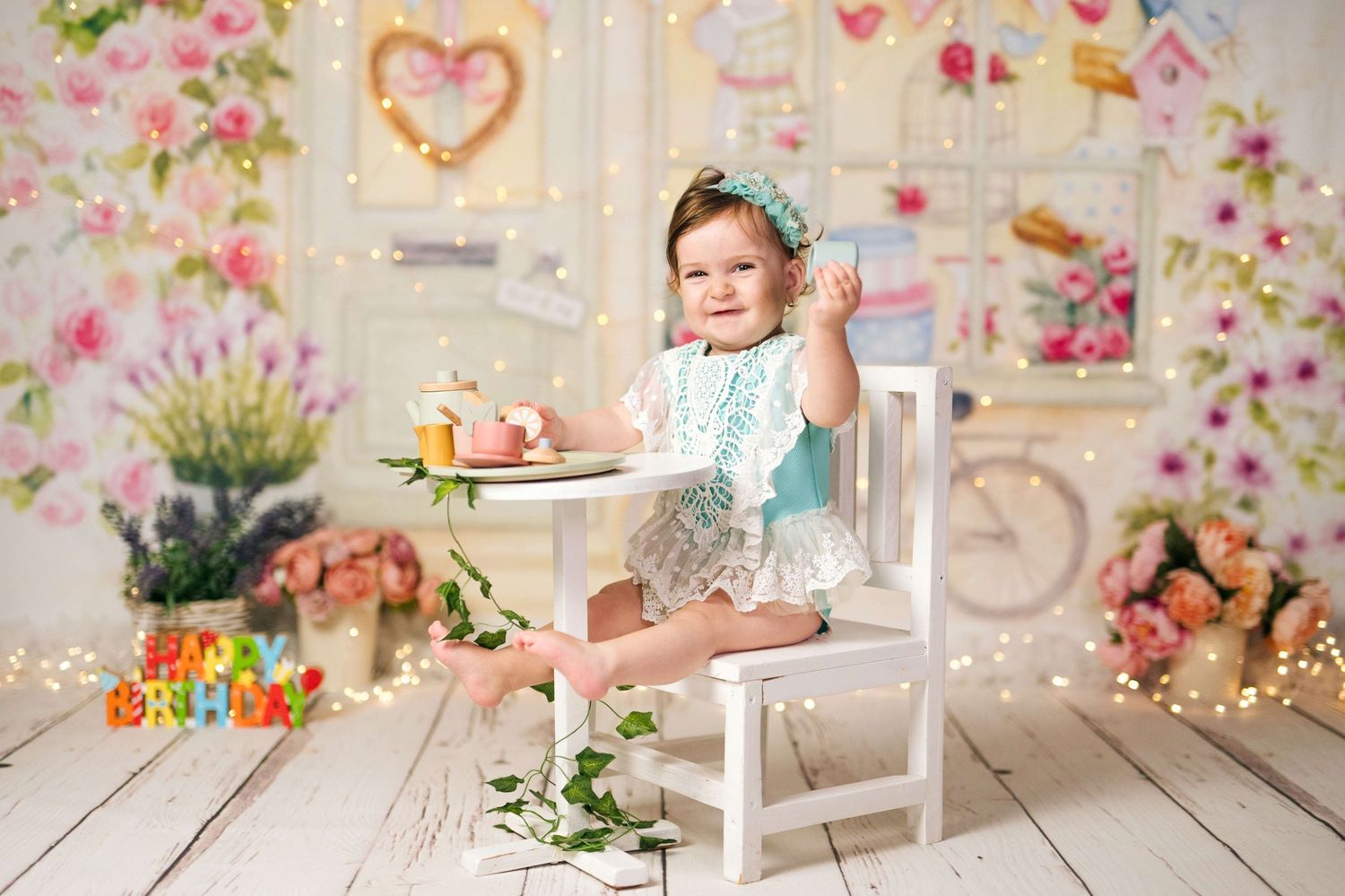 Baby girl in teal outfit celebrates first birthday with cake smash in floral decorated studio.