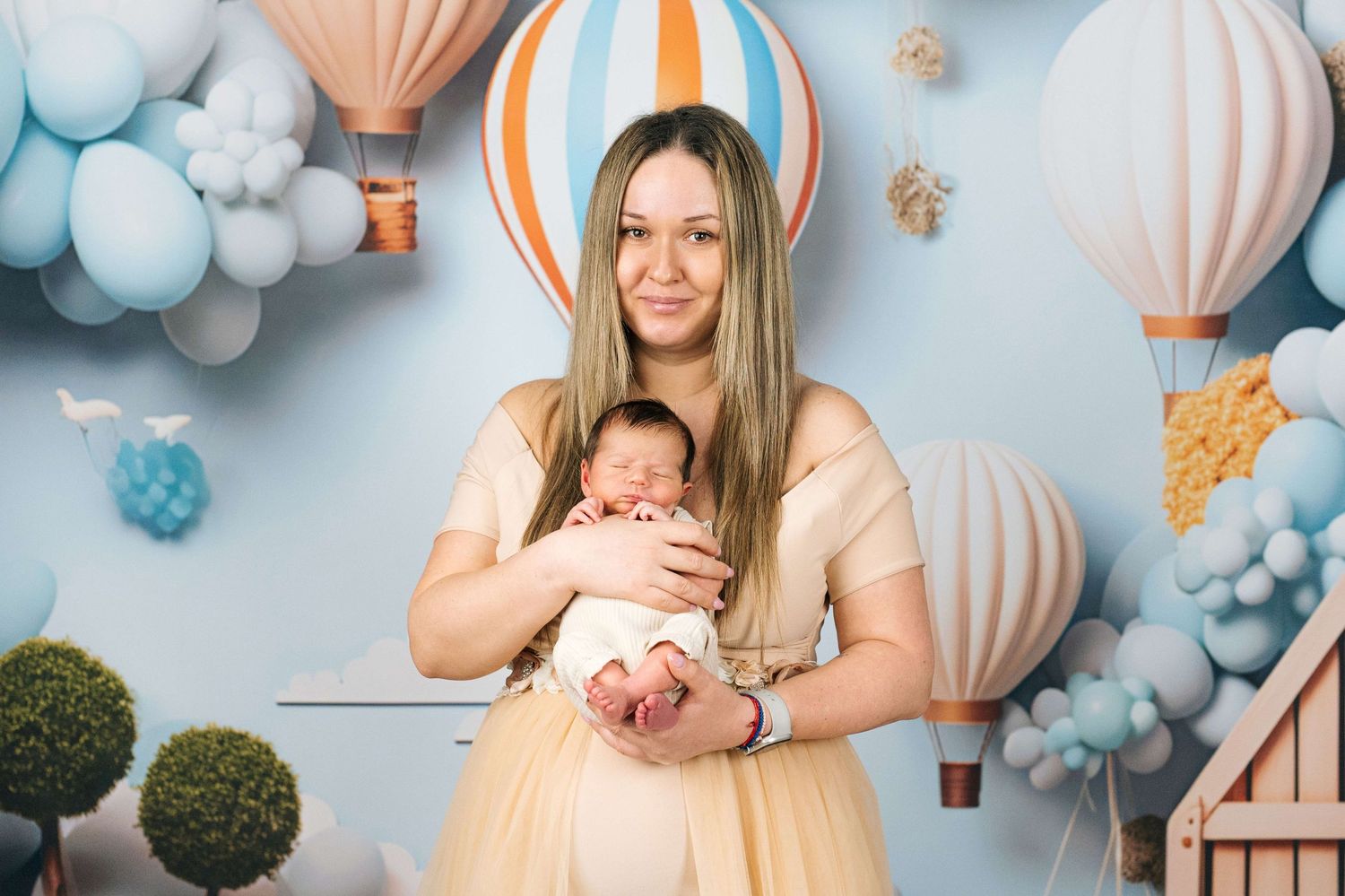A smiling woman in a gold dress holds a sleeping newborn baby against a blue backdrop with hot air balloons.