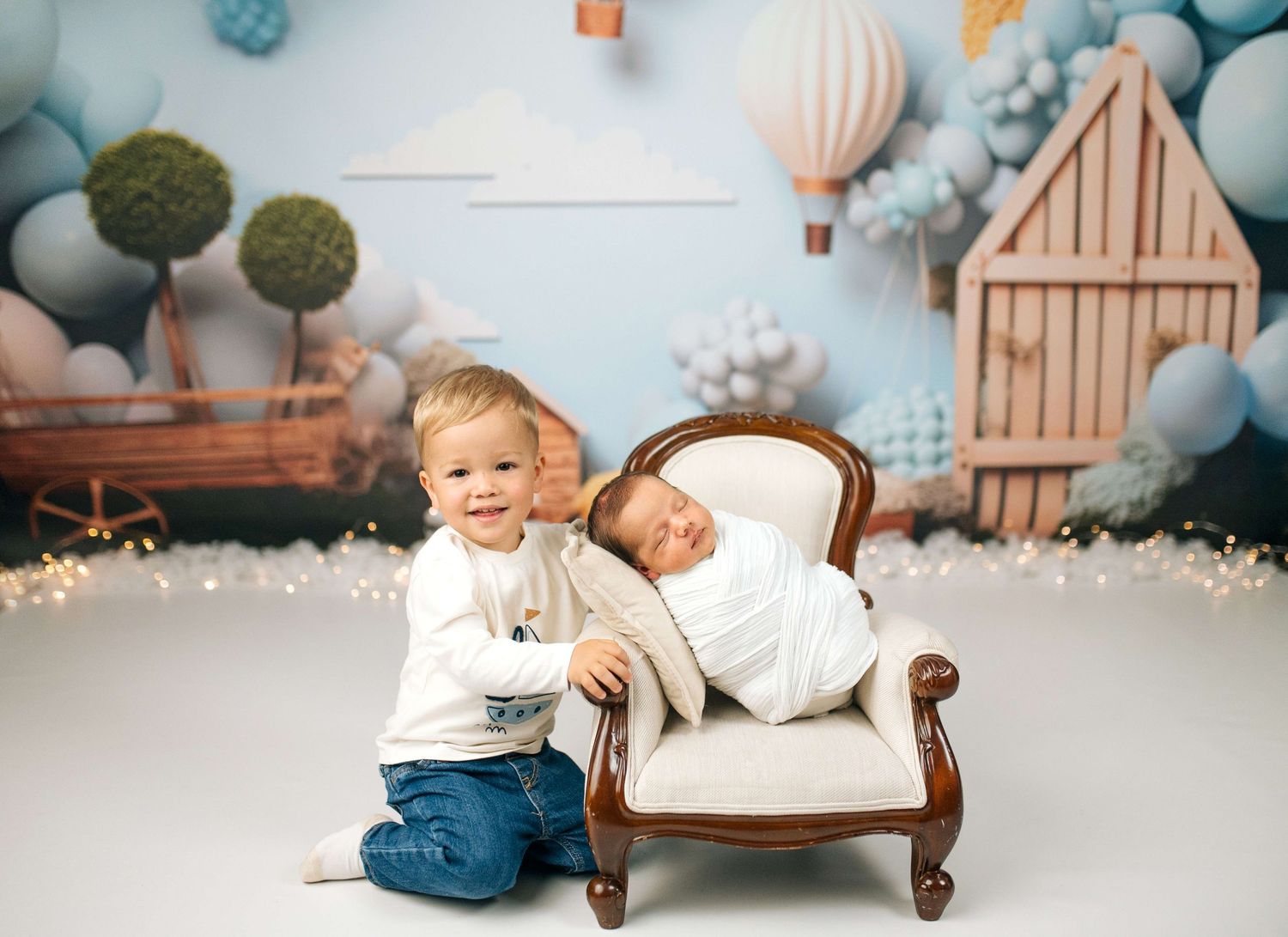 Toddler boy sits beside newborn sibling in mini chair during professional studio photo session with balloon backdrop.