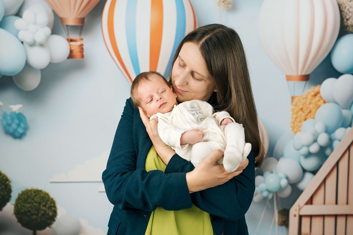 A mother tenderly holds her newborn baby in a decorated room with pastel blue balloons and hot air balloon backdrop.