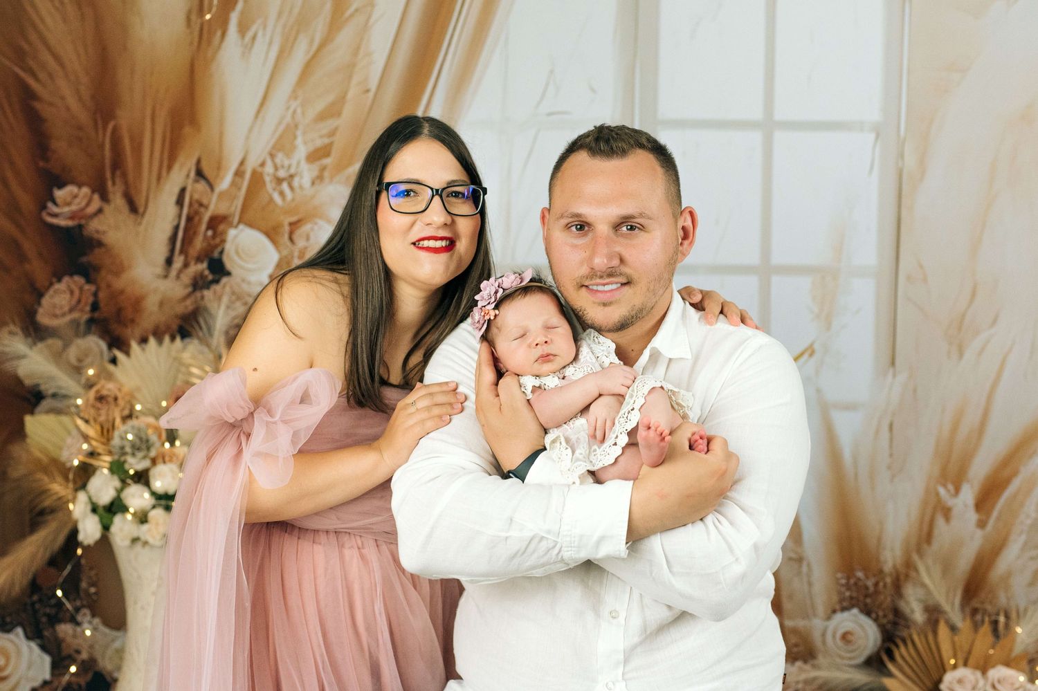 Happy family portrait with mother in pink dress, father in white shirt, and newborn baby with floral headband.